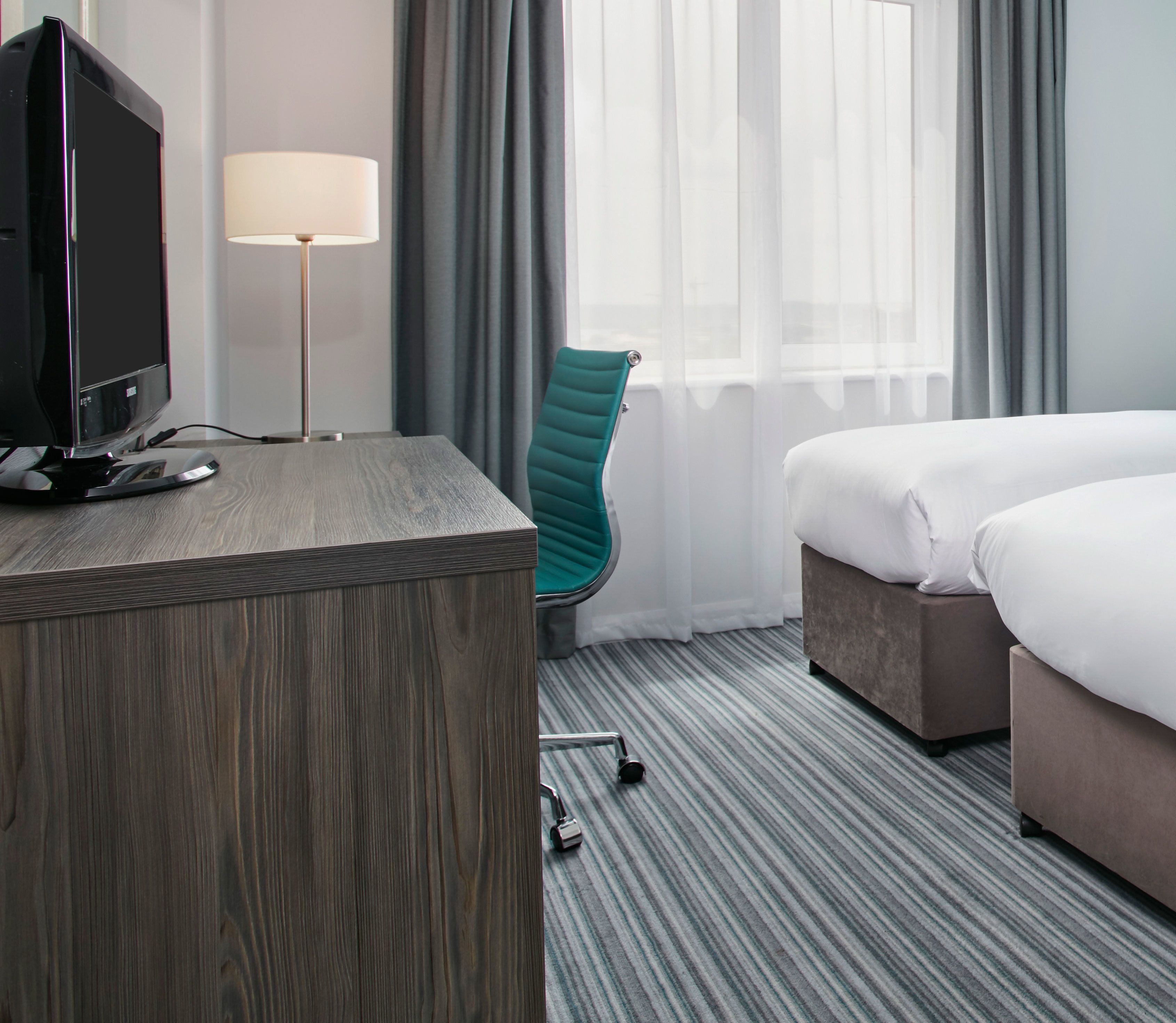 A hotel room with a wooden desk, a teal chair, a bed, and a lamp.