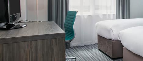 A hotel room with a wooden desk, a teal chair, a bed, and a lamp.