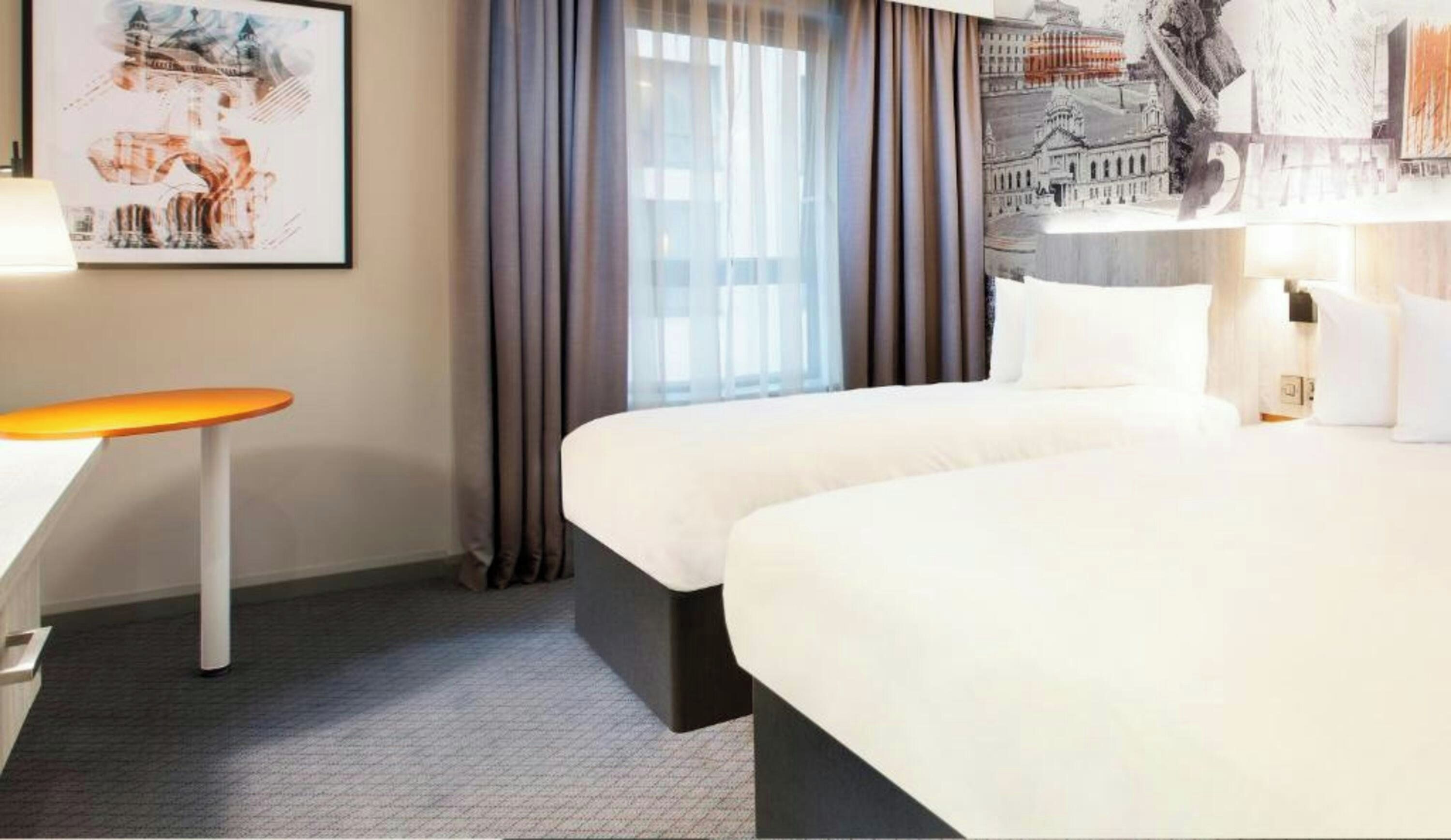A hotel room with a bed, a desk, a chair, and a large window with curtains.
