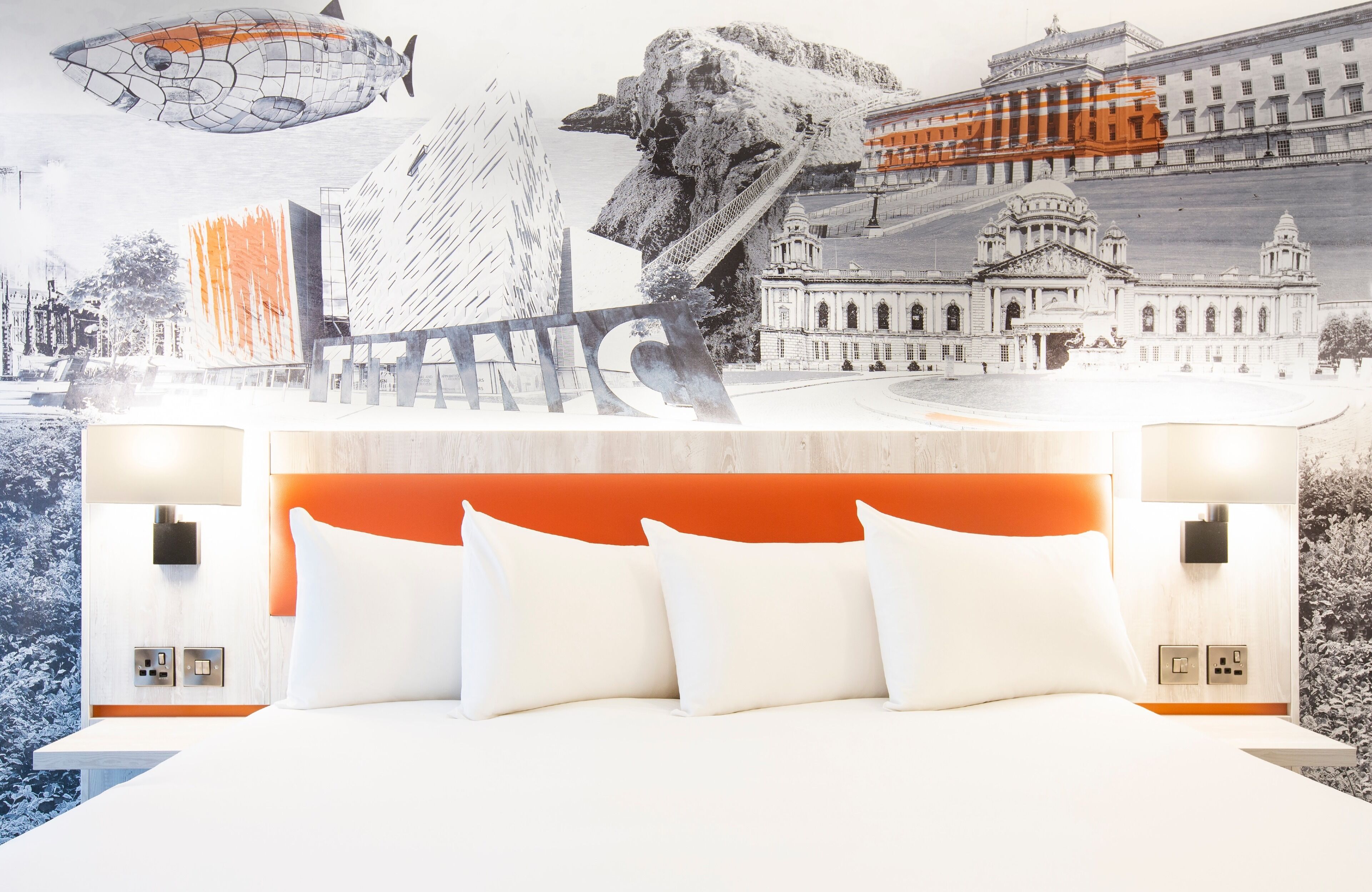 A hotel room with a bed, white pillows, an orange headboard, and a wall mural.