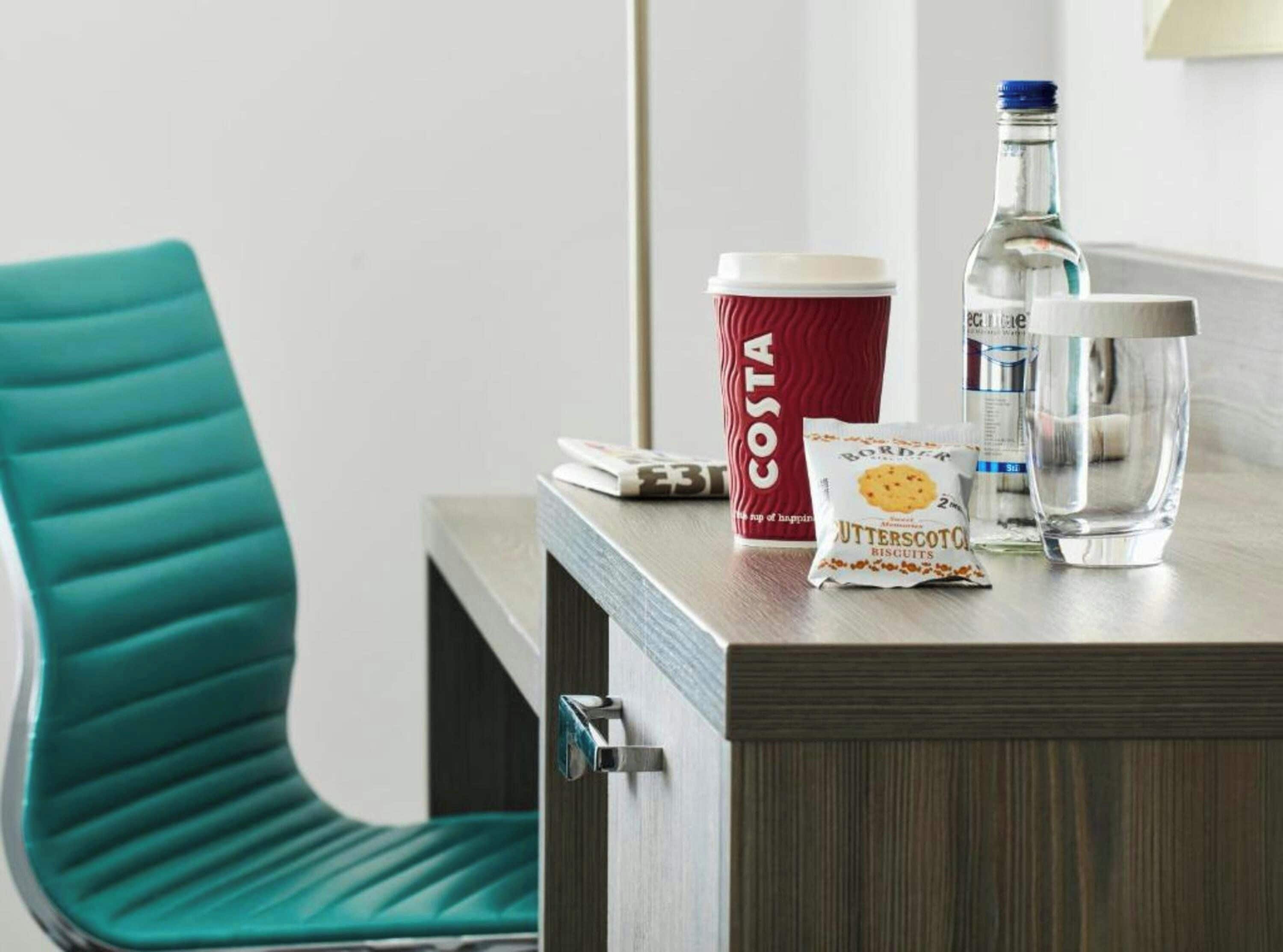 Una habitación de hotel con una silla color turquesa, una mesita de noche de madera, un vaso Costa rojo, una botella de agua, un vaso y una bolsita de galletas.