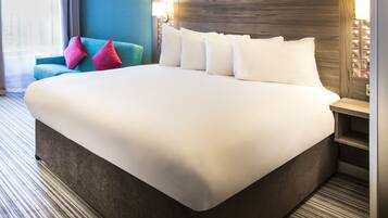 Quarto de hotel com uma cama grande, cabeceira com luminária, poltrona azul-turquesa e mesa de cabeceira com abajur.