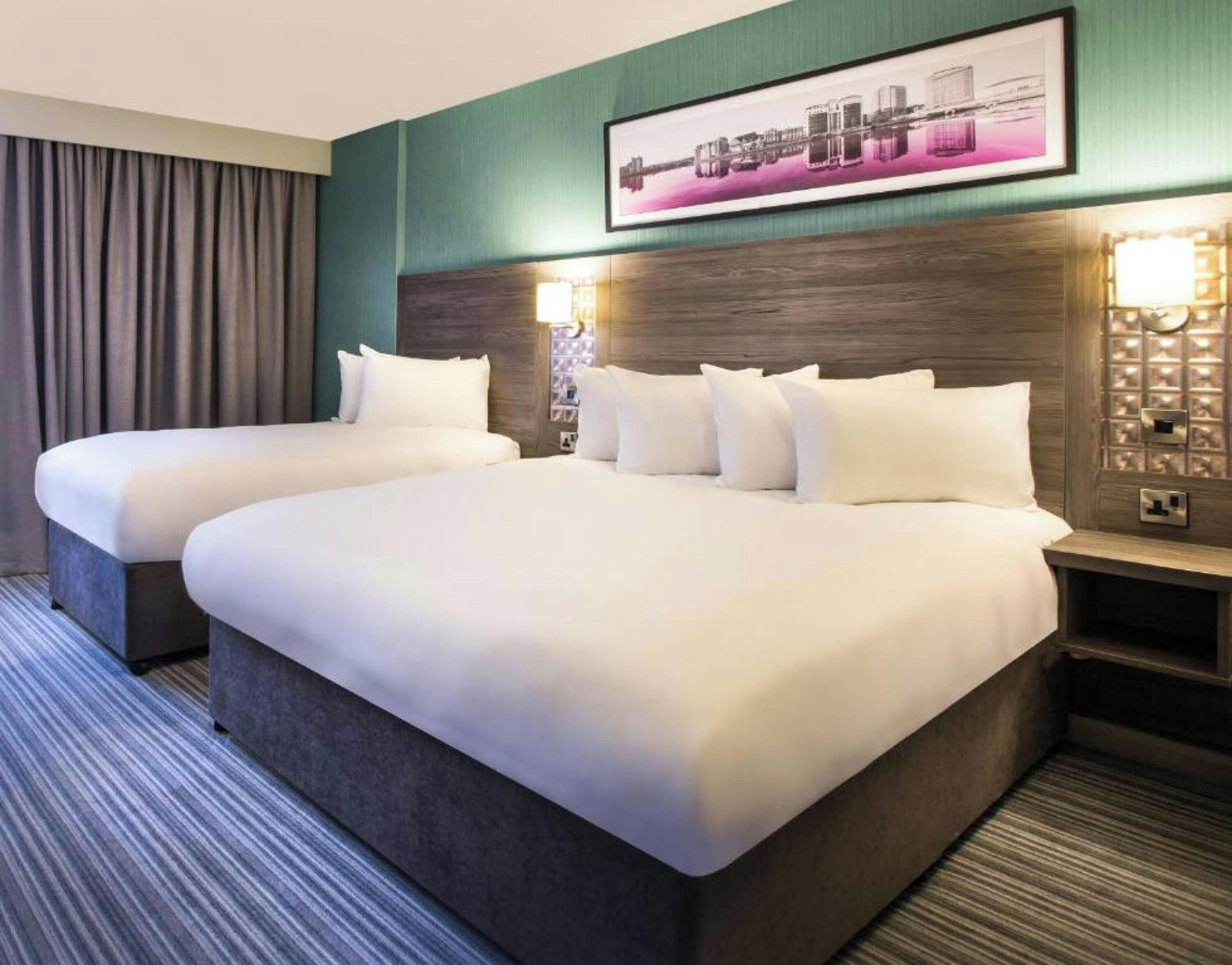 Habitación de hotel con dos camas, un cuadro grande sobre la cabecera y una mesita de noche con una lámpara.