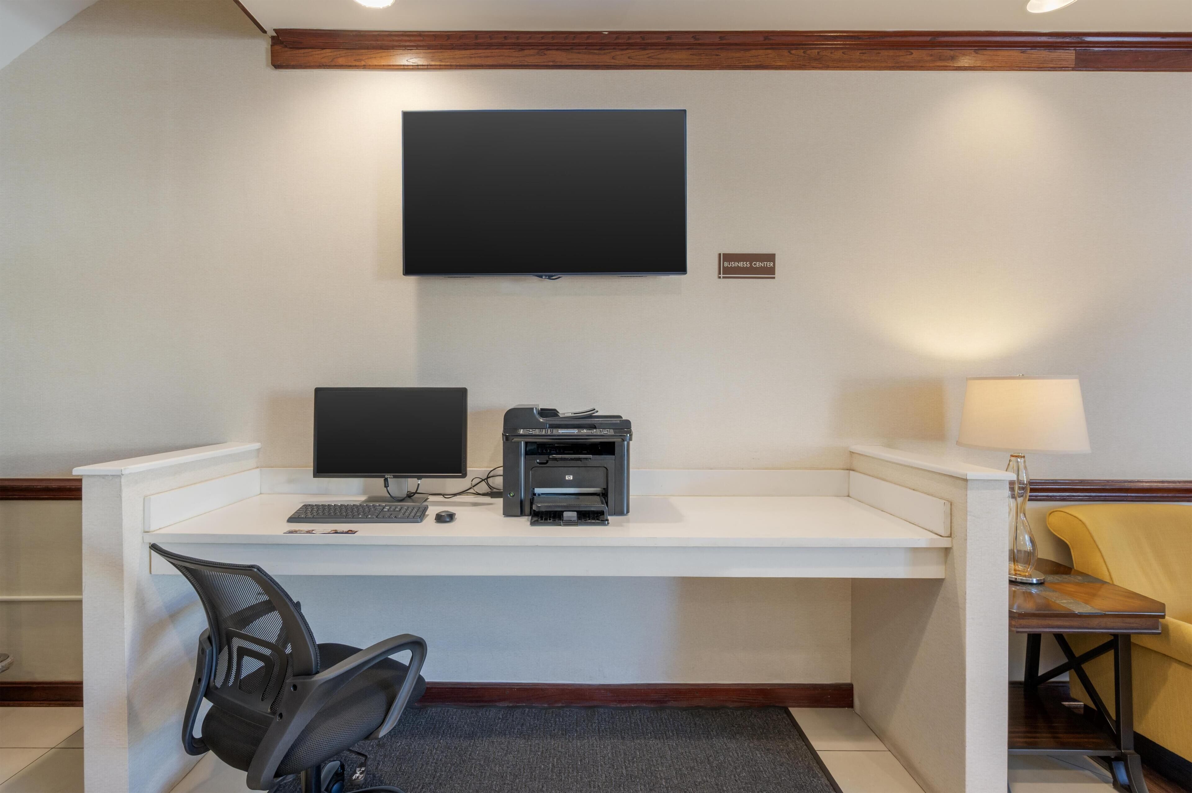A hotel room with a desk, a computer, a printer, a TV mounted on the wall, and a lamp.