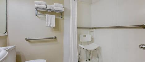 Un baño con inodoro, barandilla, ducha con barra de apoyo y una silla blanca.