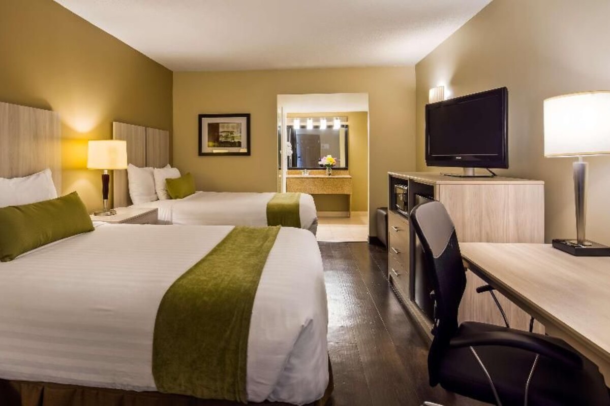 A hotel room with two beds, a desk, a television, and a bathroom with a mirror and lights.
