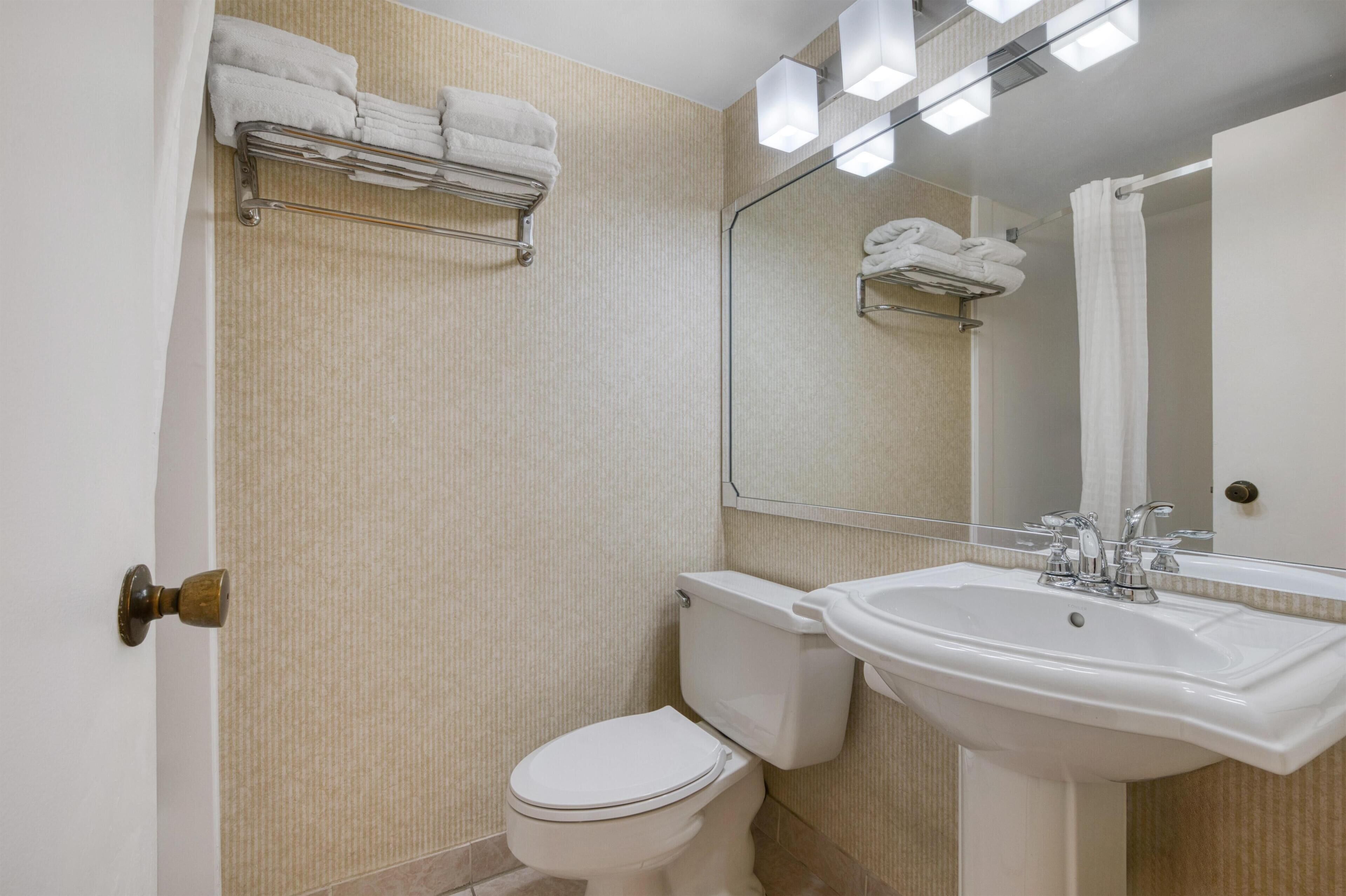 Une salle de bain avec des toilettes blanches, un lavabo et un miroir. Des serviettes sont posées sur un porte-serviettes au-dessus des toilettes.