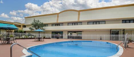 Una piscina con cerca blanca, un área de patio techada y un edificio de techo plano al fondo.