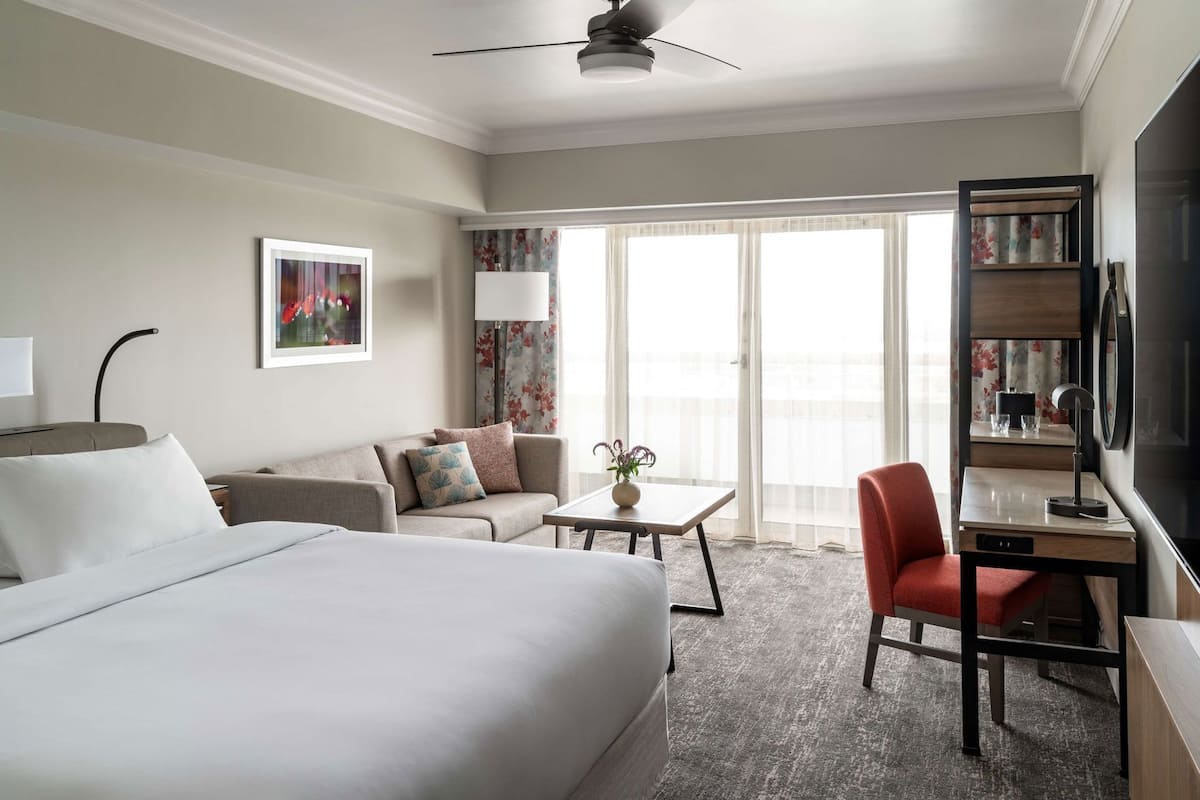 Habitación de hotel con cama, sofá, escritorio, silla, televisor y balcón con cortinas.
