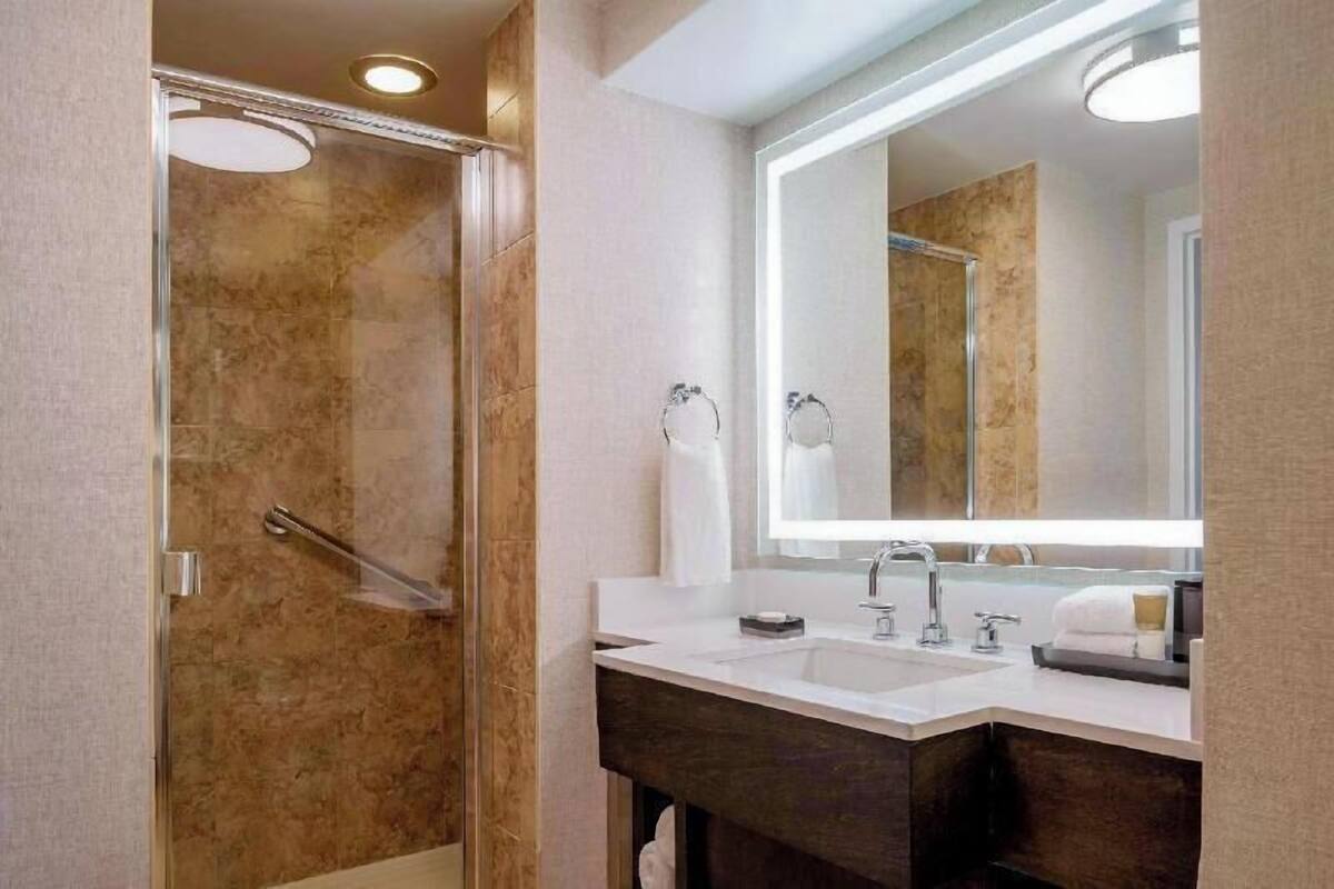 Un baño moderno con ducha envuelta en vidrio, un espejo grande y un tocador con encimera oscura.