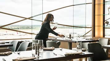Un espace repas moderne, avec des tables dressées pour le repas, de grandes fenêtres offrant une vue sur l’aéroport, et une personne s’occupant de la table.