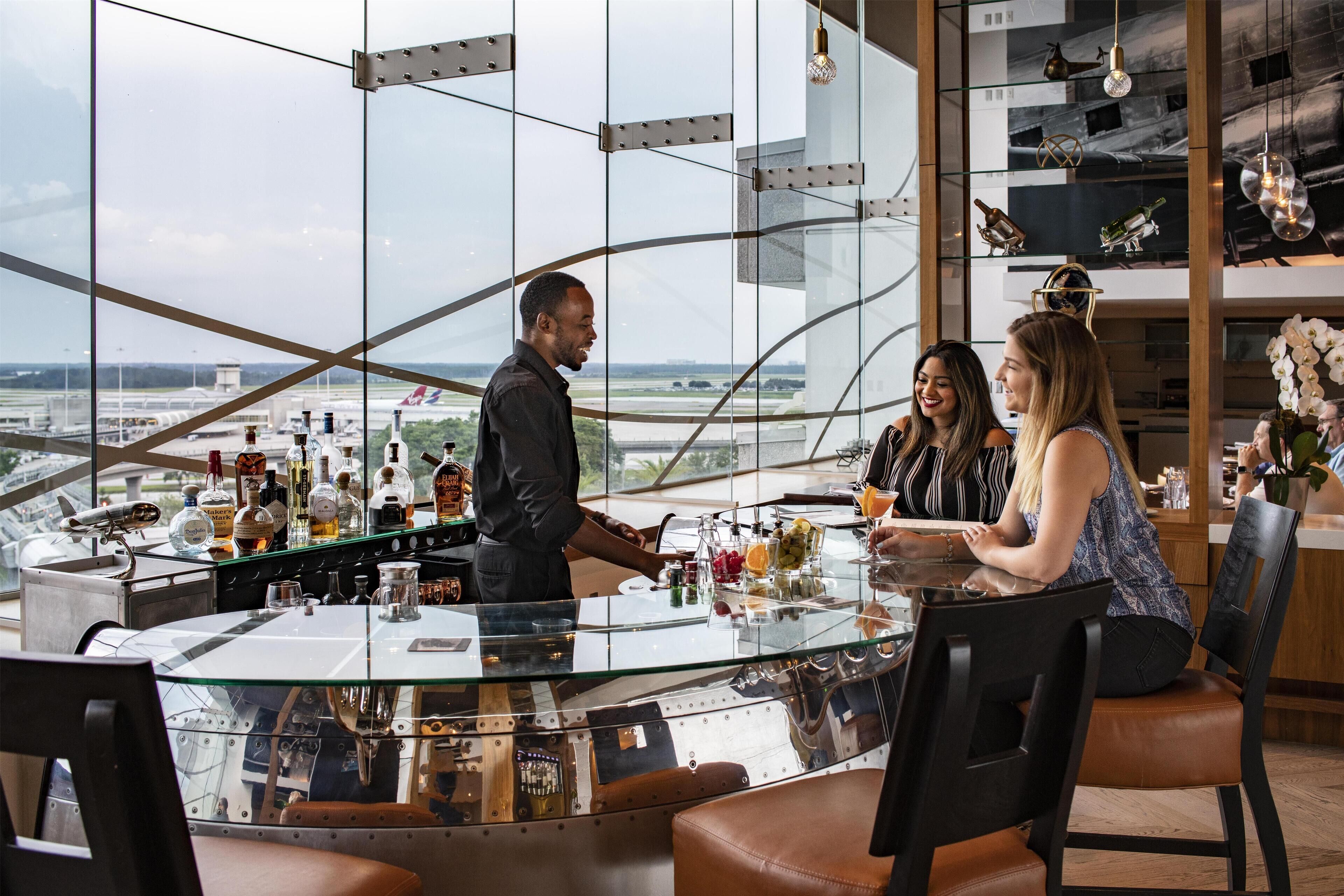 A modern bar with a glass counter, various bottles, and a view of an airport.