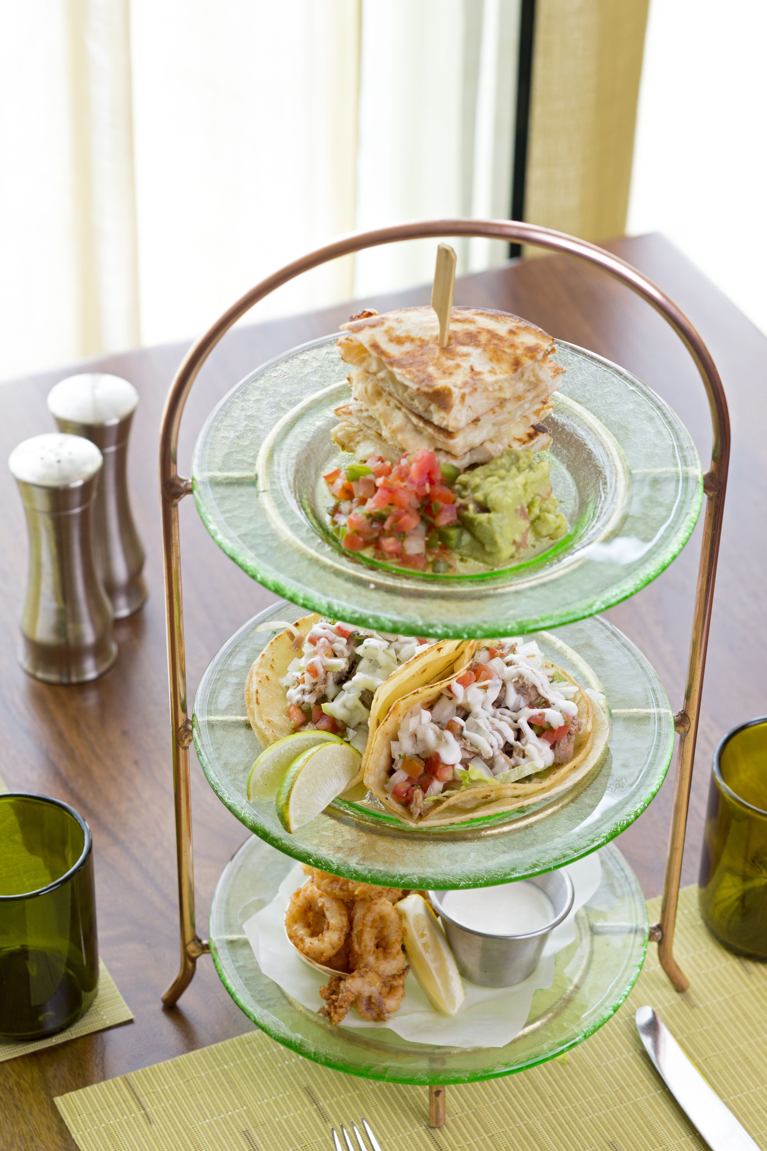 Una bandeja de vidrio de tres niveles con diversos platillos, entre ellos tacos, guacamole y mariscos.