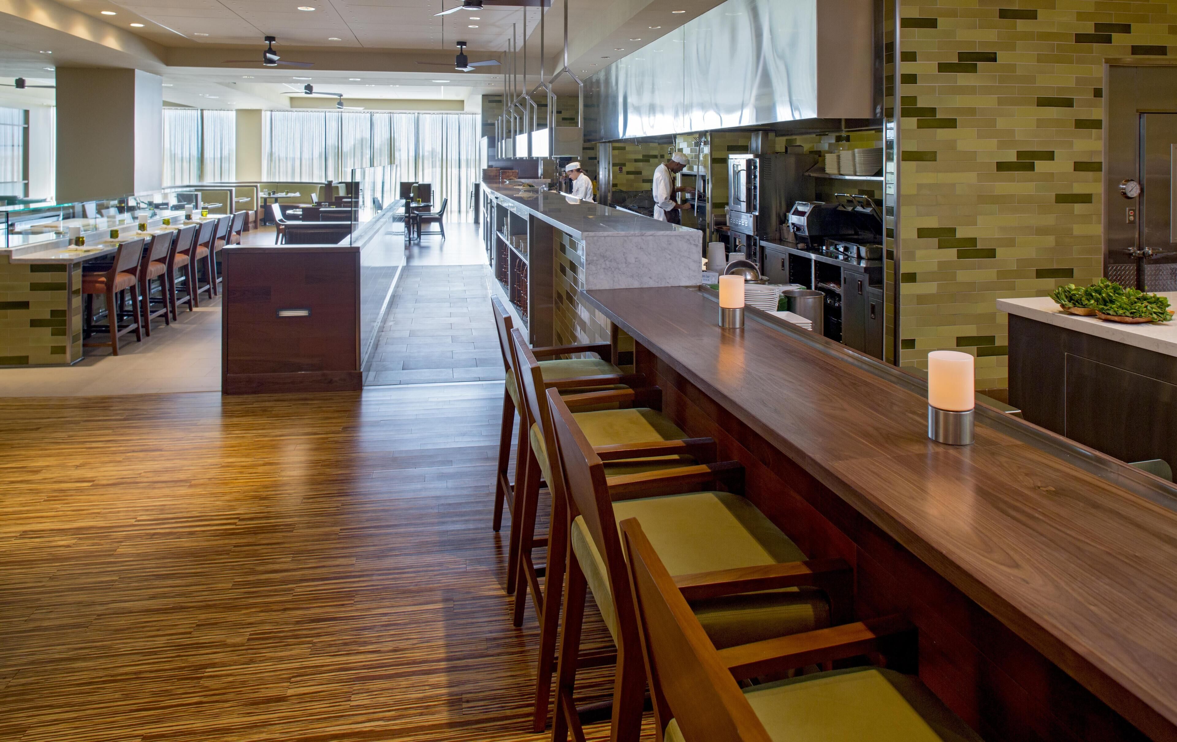 A modern restaurant with a long counter, bar seating, and a kitchen area.