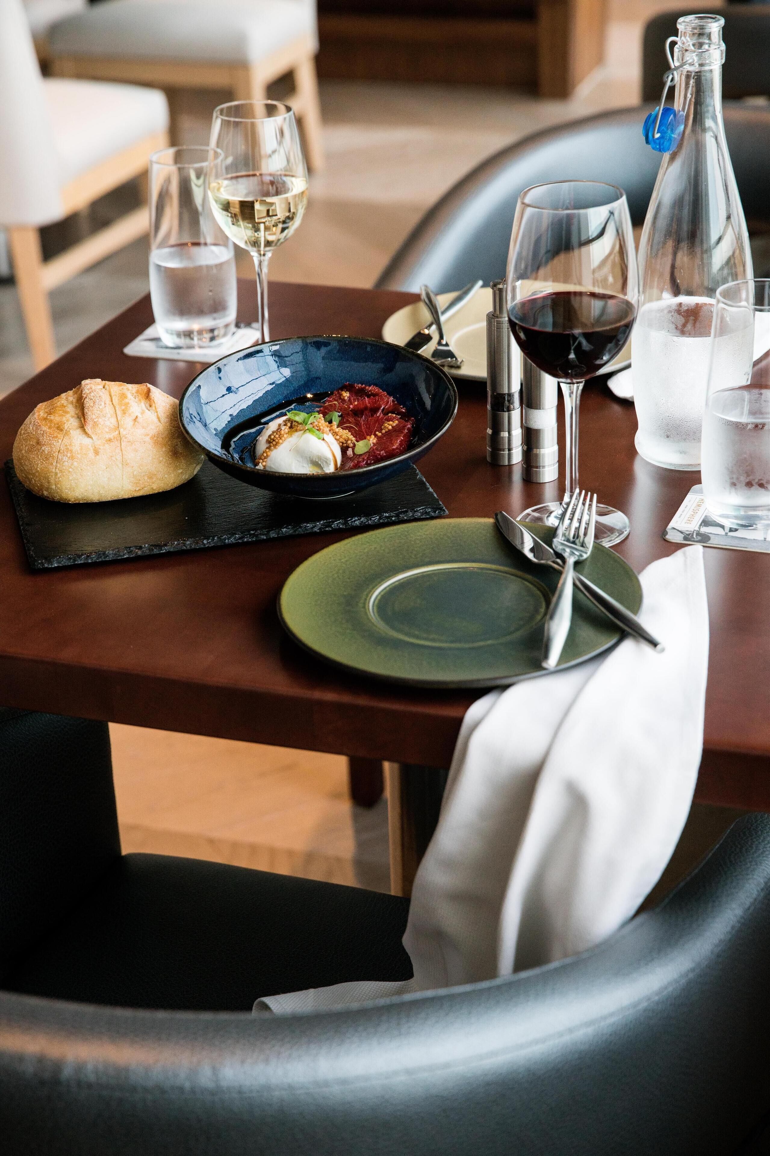 Una mesa de comedor dispuesta con un plato, cubiertos, un panecillo, un bol de comida y vasos de agua y vino.