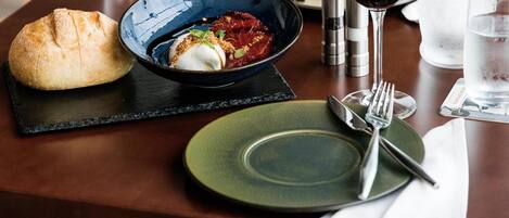 Una mesa de comedor dispuesta con un plato, cubiertos, un panecillo, un bol de comida y vasos de agua y vino.