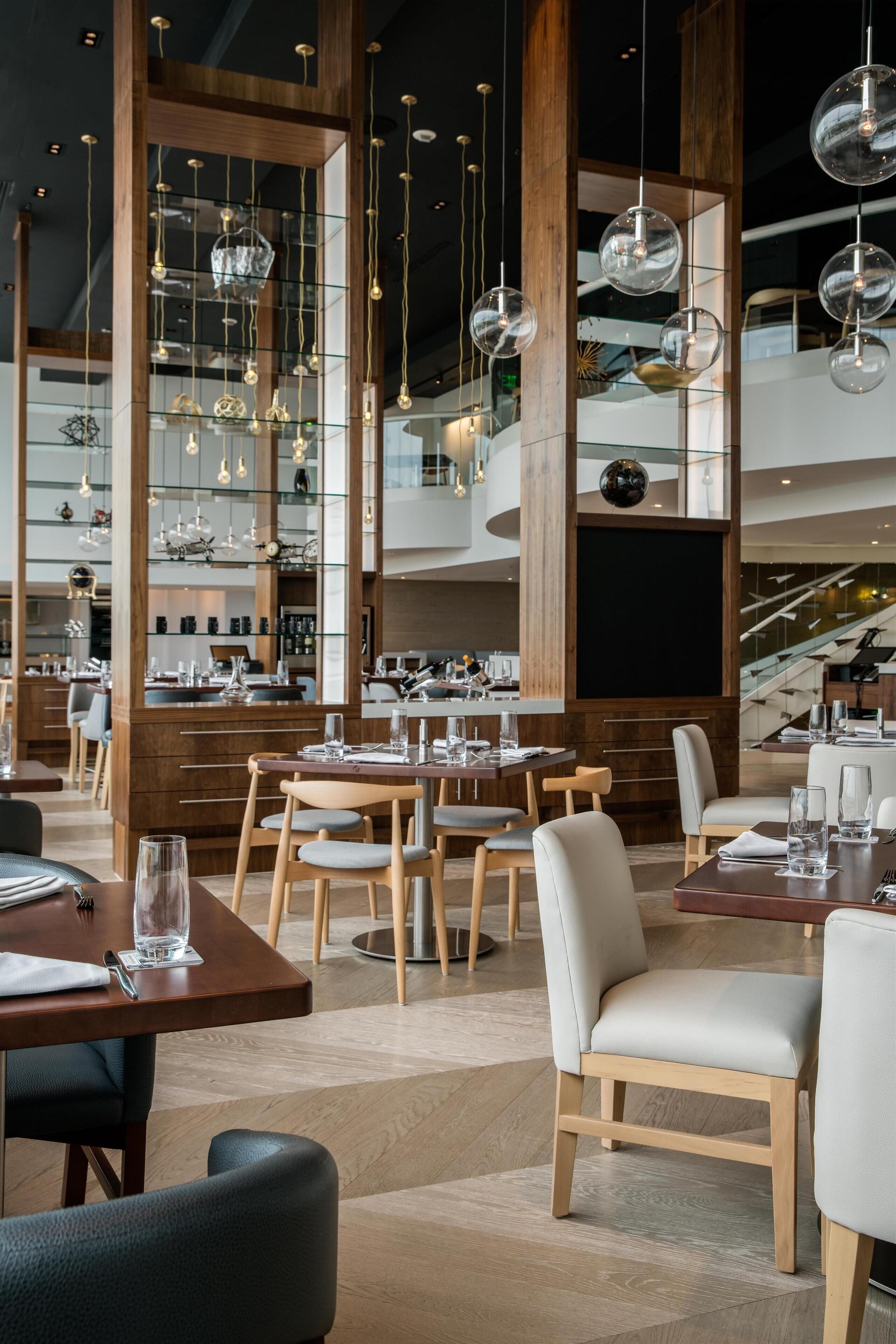 A modern restaurant interior with high ceilings, wooden beams, and hanging spherical light fixtures. Tables are set with white tablecloths and chairs.