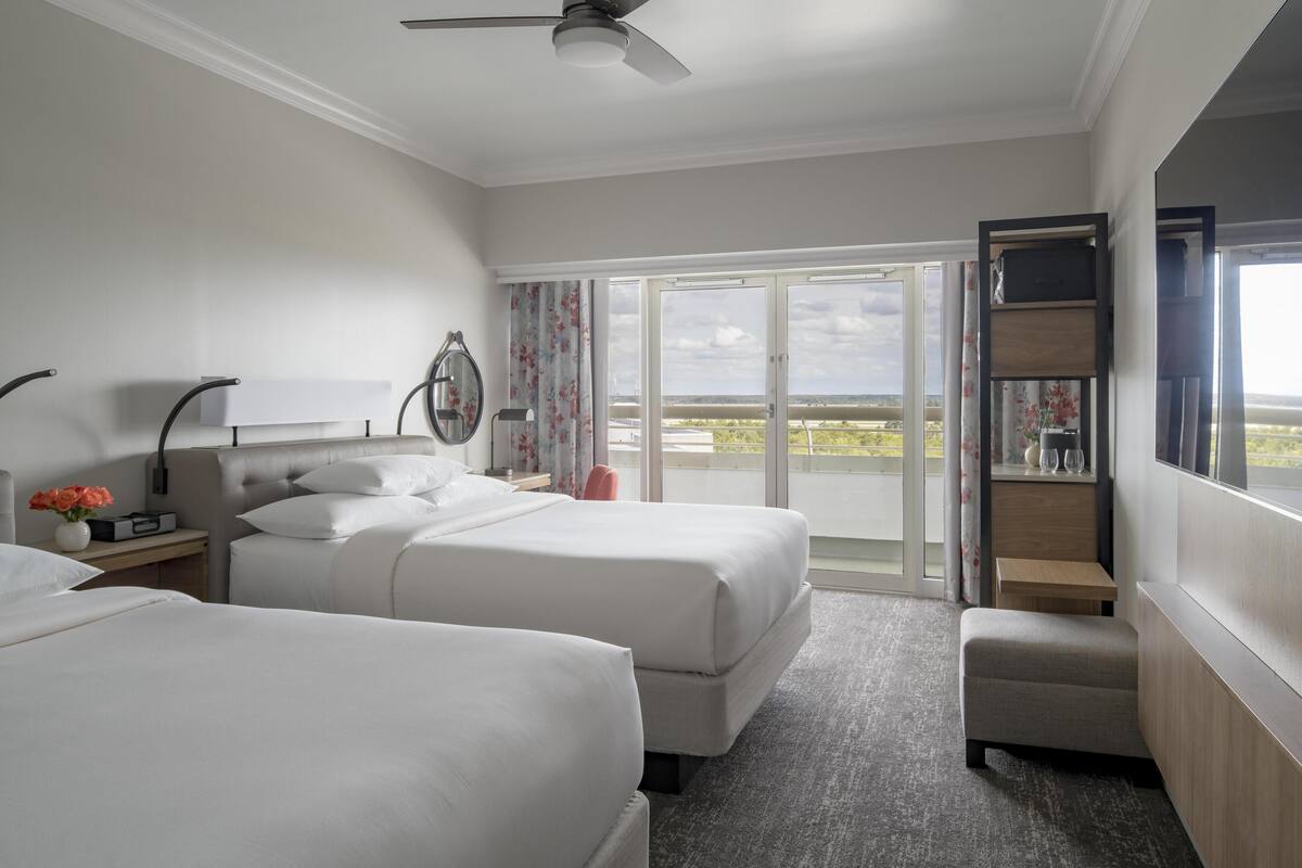 Habitación de hotel con dos camas, una mesita pequeña, una silla, un ventilador de techo y vistas al exterior a través de un ventanal corredero.
