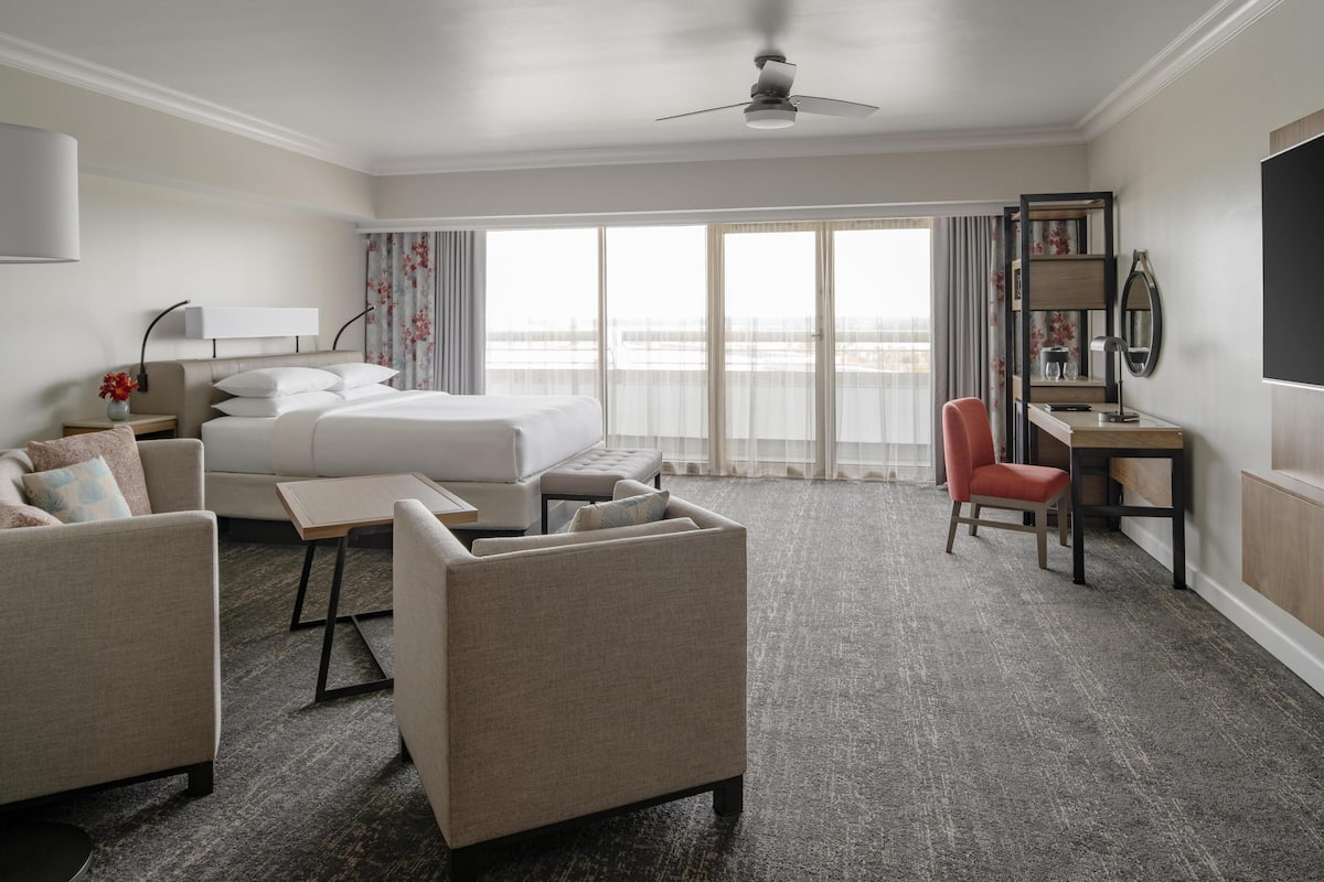 Habitación de hotel con cama, dos sillones, escritorio, silla, televisor y balcón con cortinas.