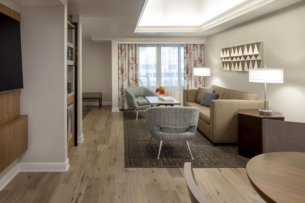 Una habitación de hotel moderna con piso de madera, un sofá, una silla, una mesita y una lámpara.