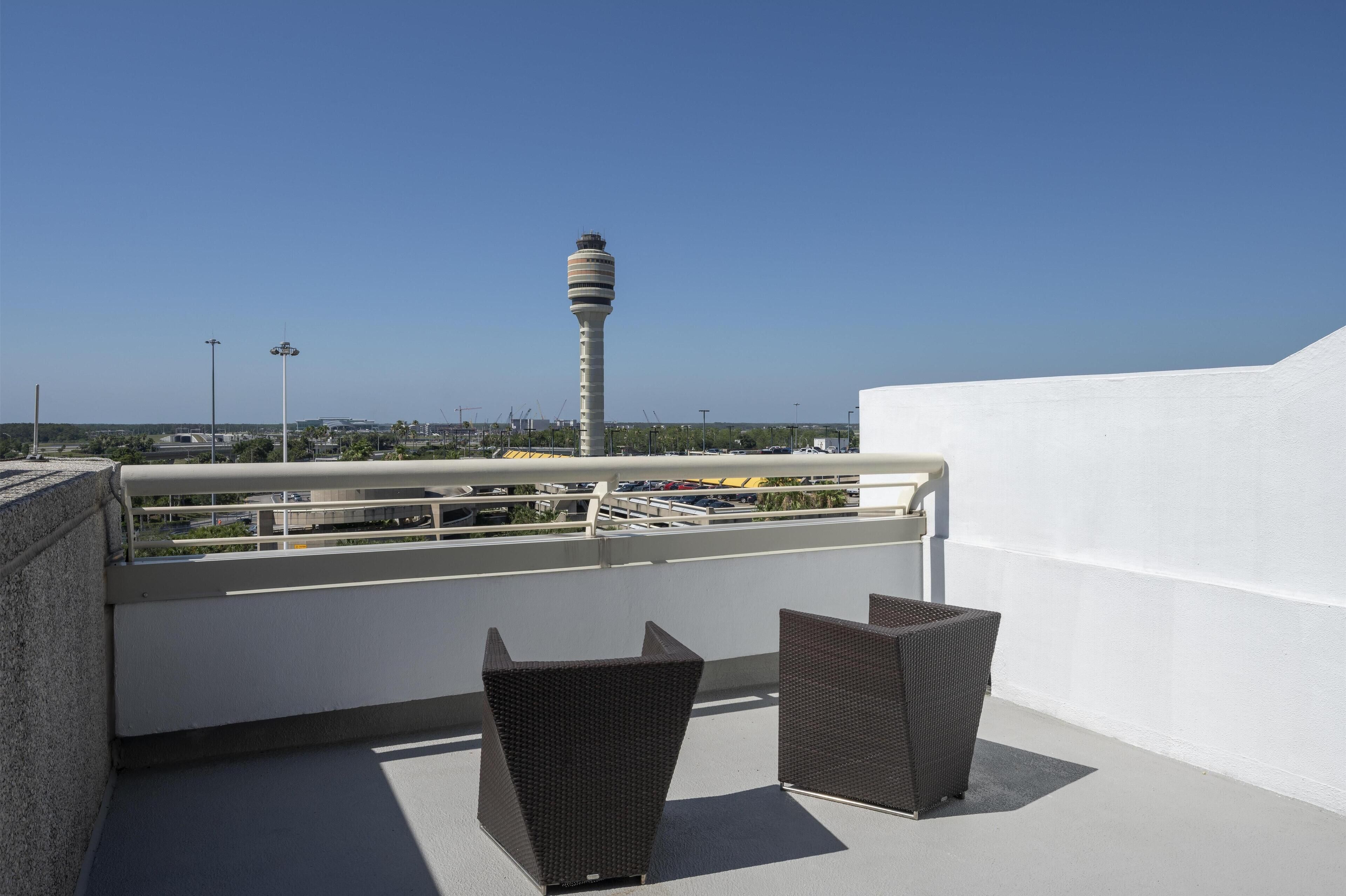 Terraza en la azotea con dos sillas de mimbre oscuro, una pared blanca y vistas a una torre alta y edificios a la distancia.