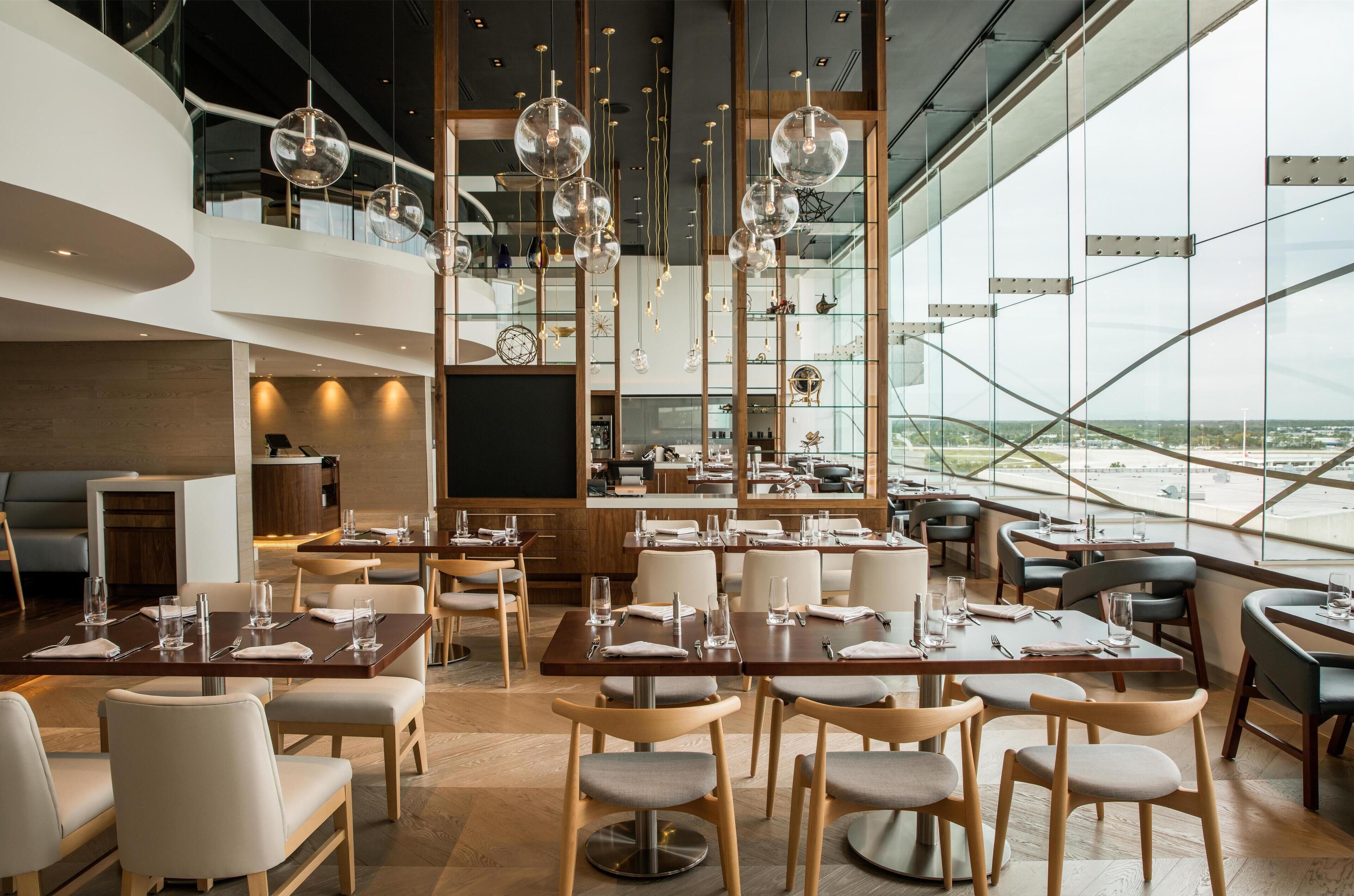 A modern restaurant with high ceilings, large windows, and a view of the city. Tables are set with white tablecloths and silverware.