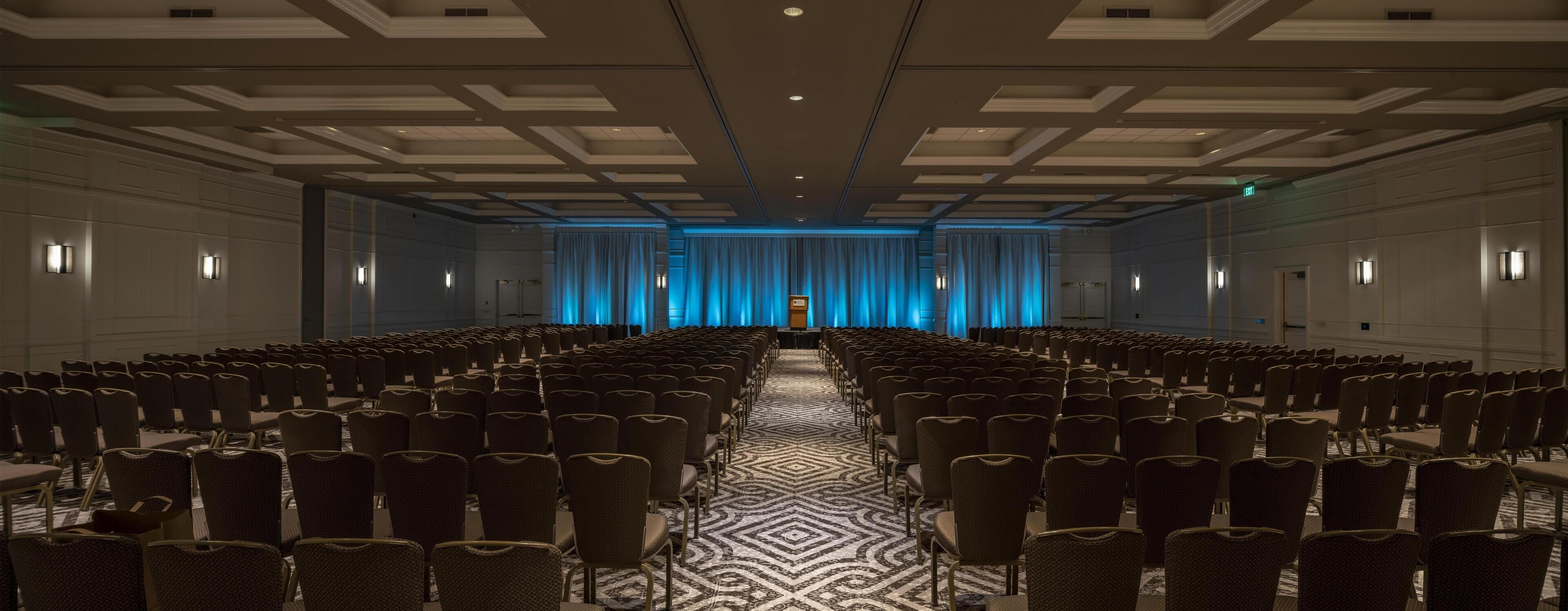 A conference room with rows of chairs, a patterned carpet, and a stage with blue curtains.
