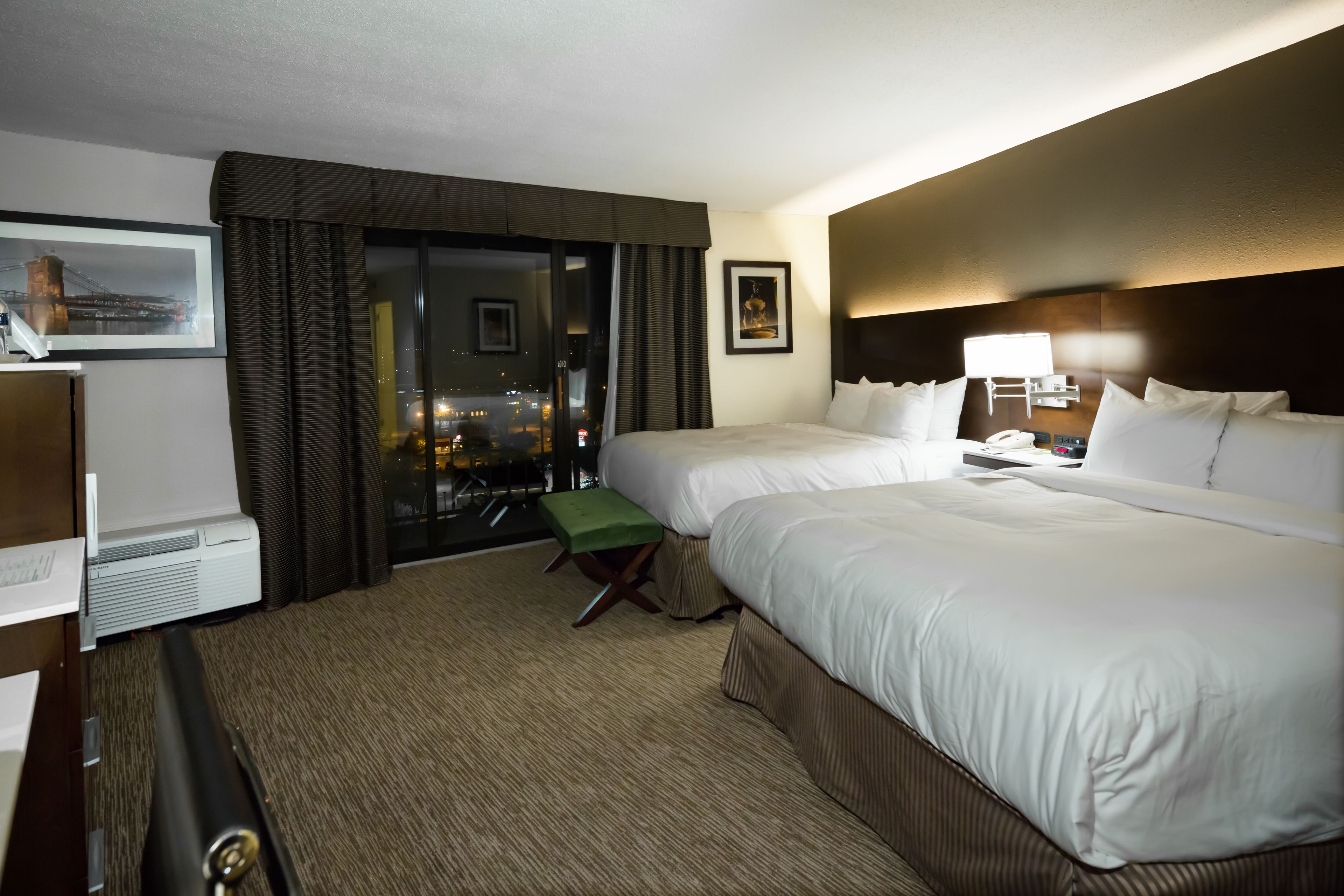 A hotel room with two beds, a desk, a chair, a large window with a view of a cityscape, and a framed picture on the wall.