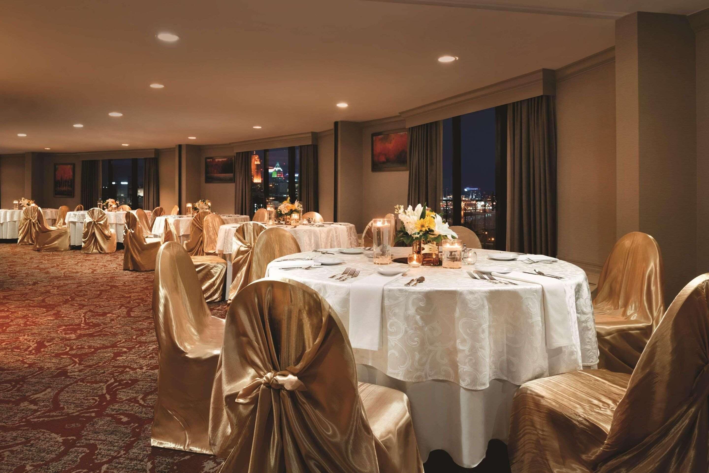 A banquet hall with round tables set for dining, covered with white tablecloths and gold chair covers, featuring a cityscape view through large windows.