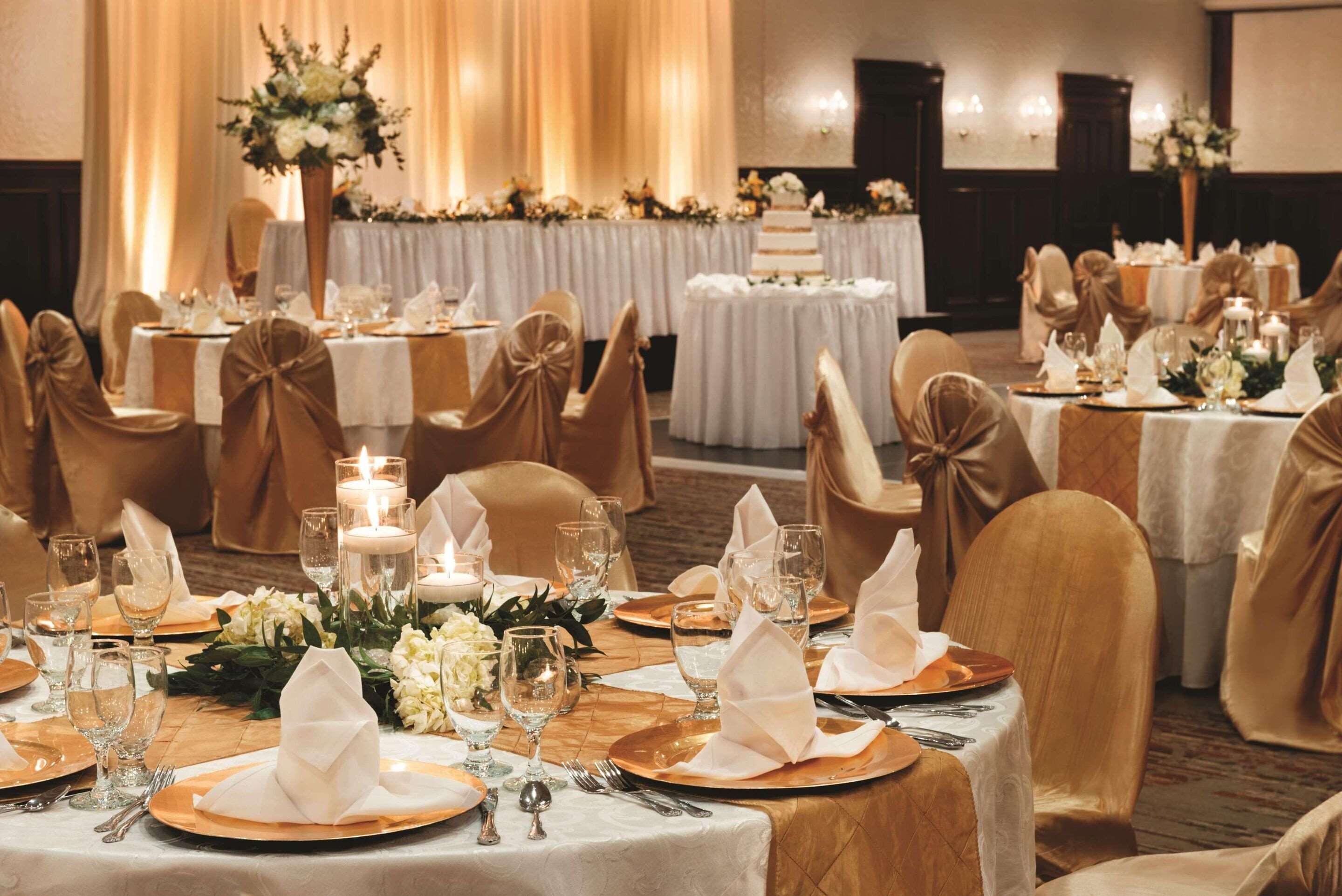 Un salón de banquetes con mesas redondas dispuestas para un evento formal, con manteles blancos, sillas doradas y centros de mesa florales.