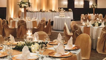 Un gran saló de banquets amb taules rodones preparades per a un esdeveniment formal, amb teles de taula blanques, cadiretes daurades i centrals florals.