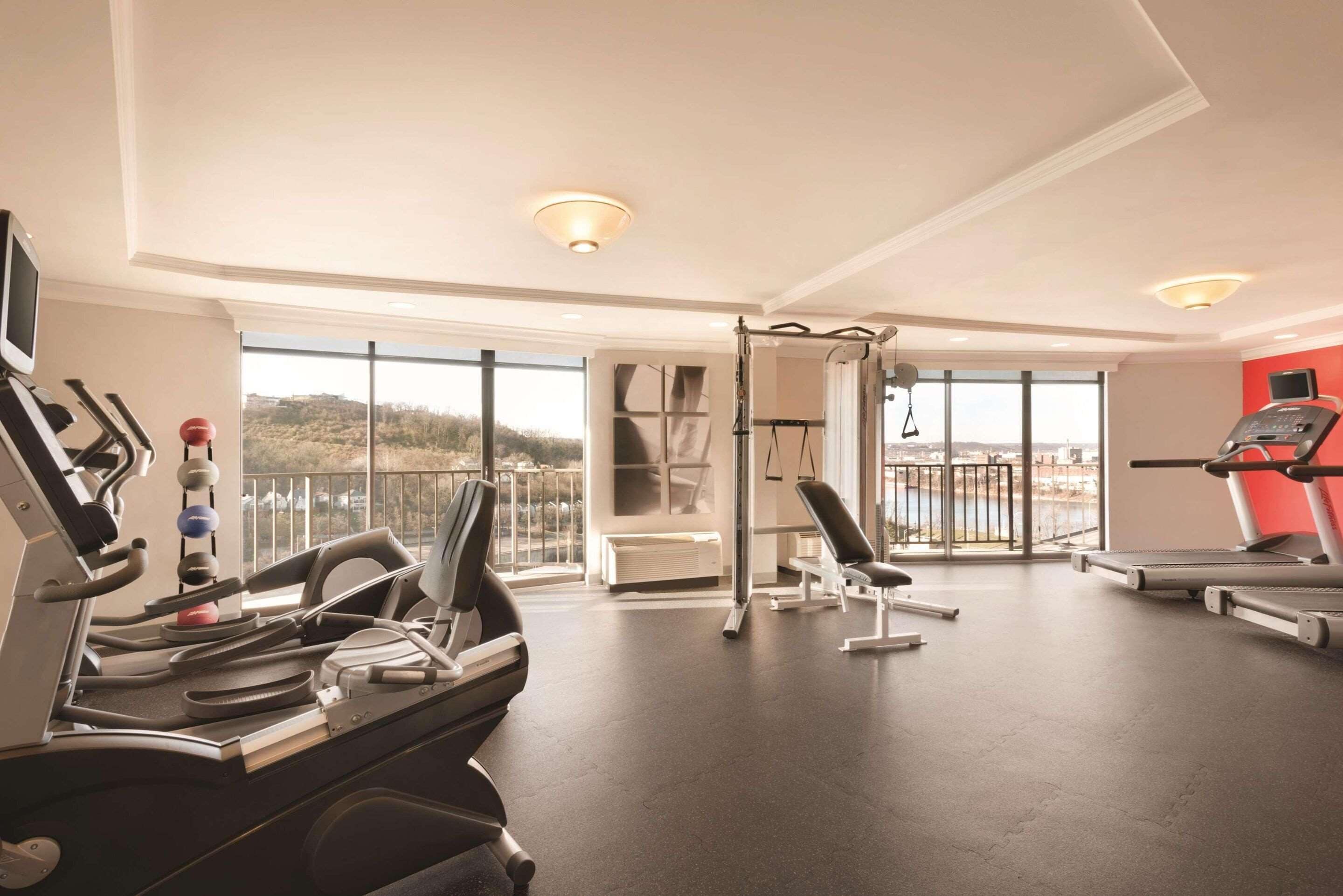 Een sportschool met loopbanden, een hometrainer en een krachttrainingapparaat. Er zijn grote ramen en uitzicht op het buitengebied.