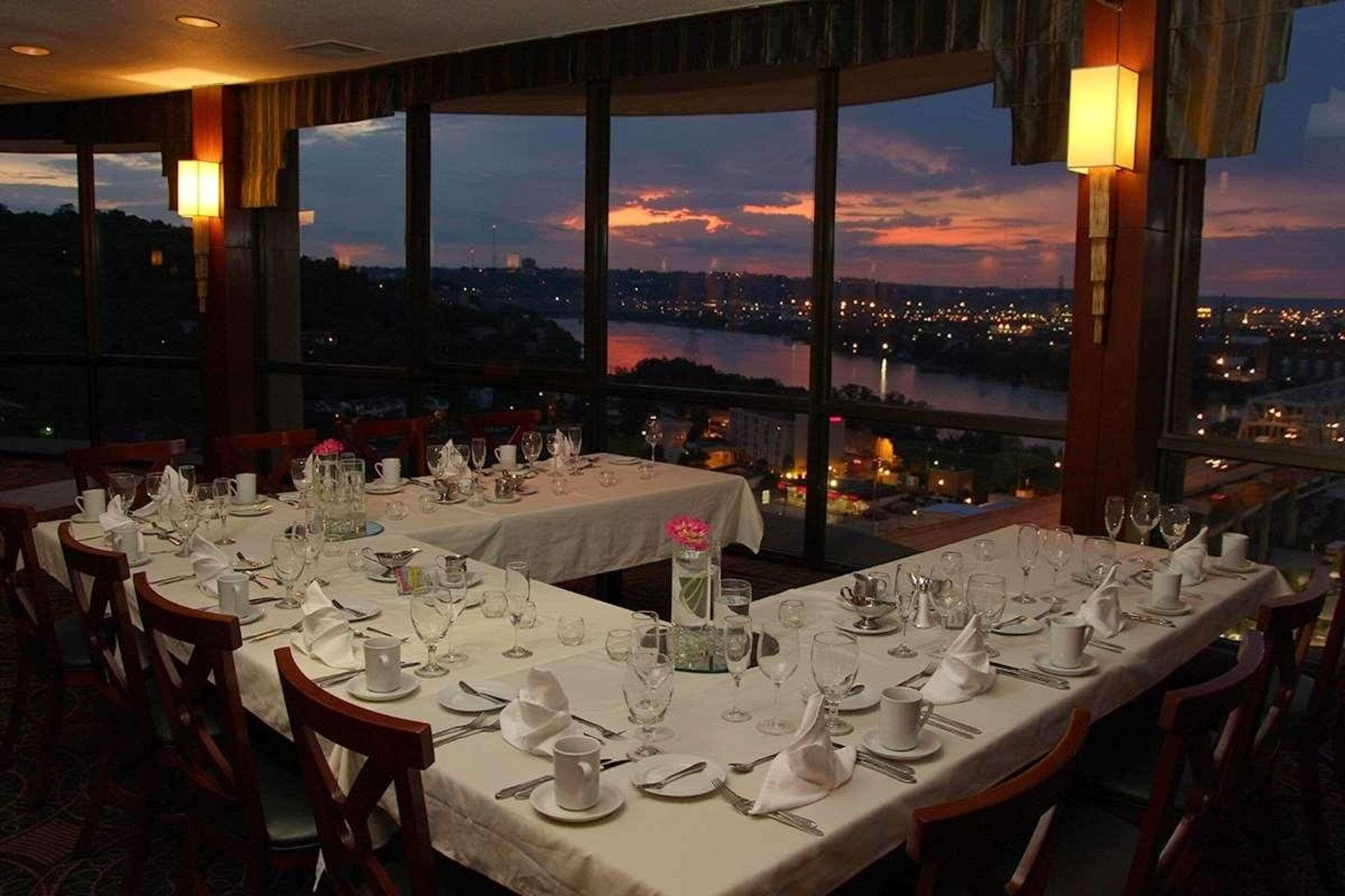 A restaurant with tables set for dining, large windows offering a view of a cityscape and a body of water at dusk.