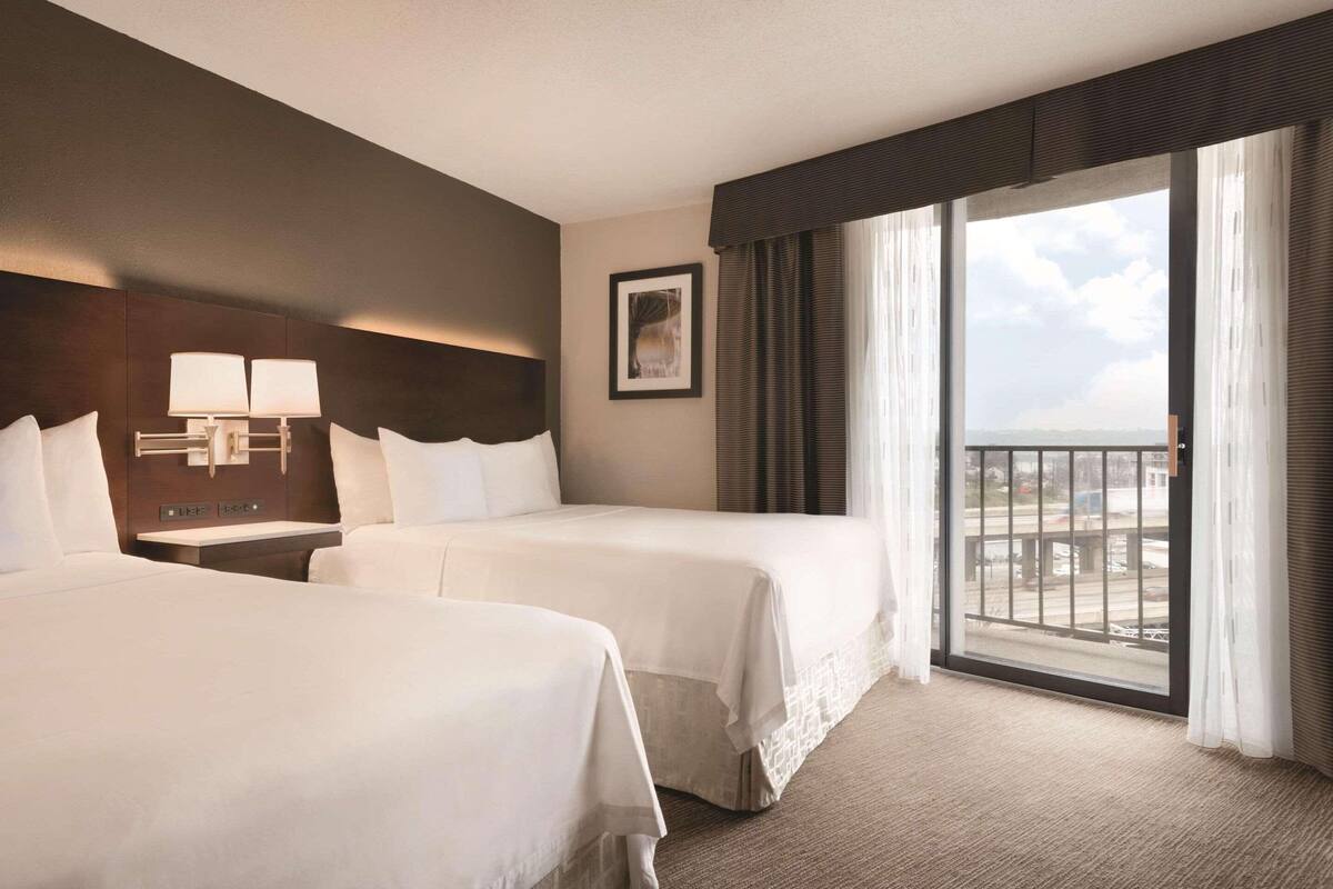 A hotel room with two beds, a large window with a view, a nightstand with lamps, and a framed picture on the wall.