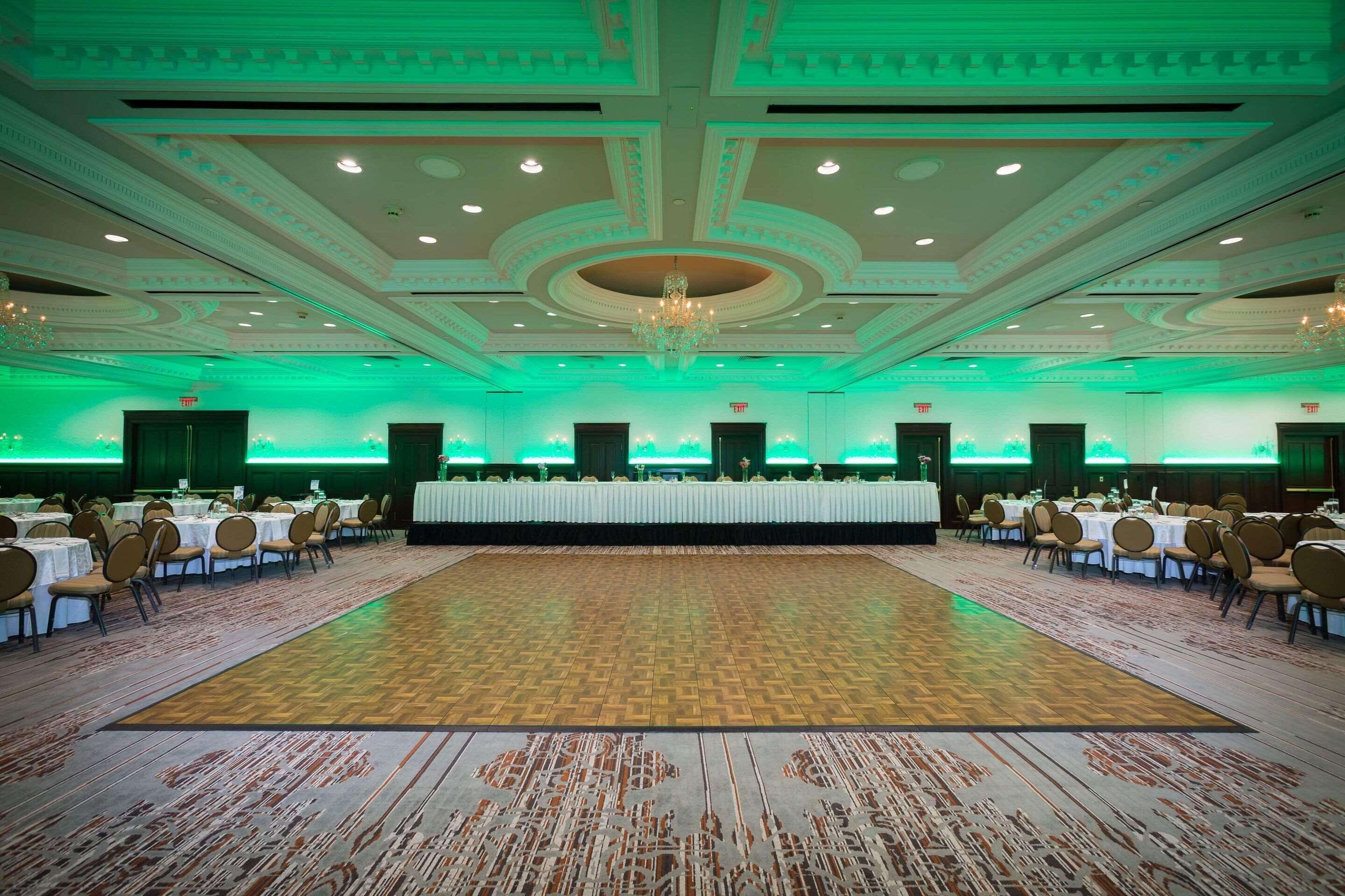 A large banquet hall with a central long table surrounded by chairs, a patterned carpet, and a chandelier.