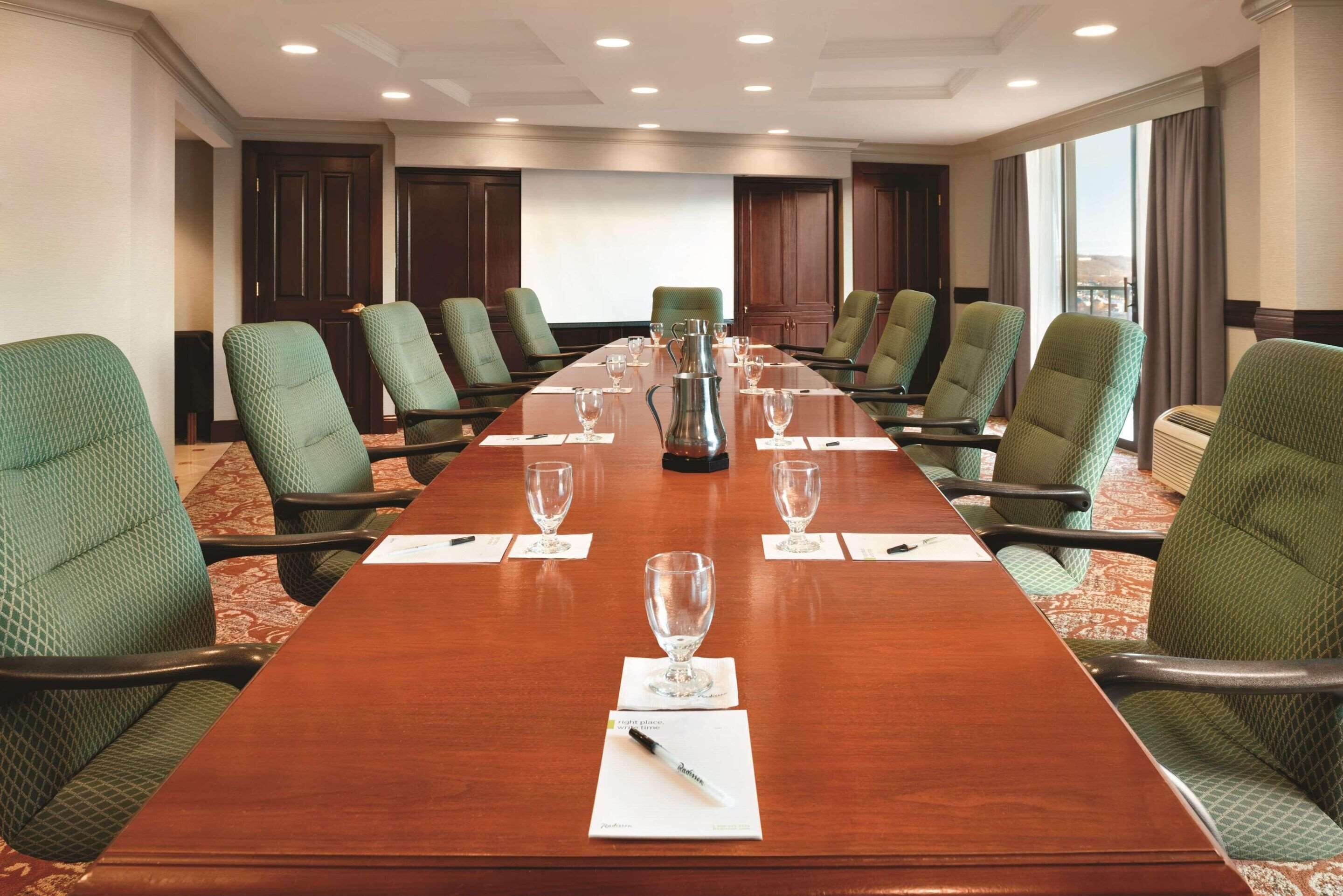 Une salle de conférence avec une longue table, des chaises recouvertes d’un tissu vert et un écran de projection.