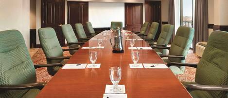 Une salle de conférence avec une longue table, des chaises recouvertes d’un tissu vert et un écran de projection.