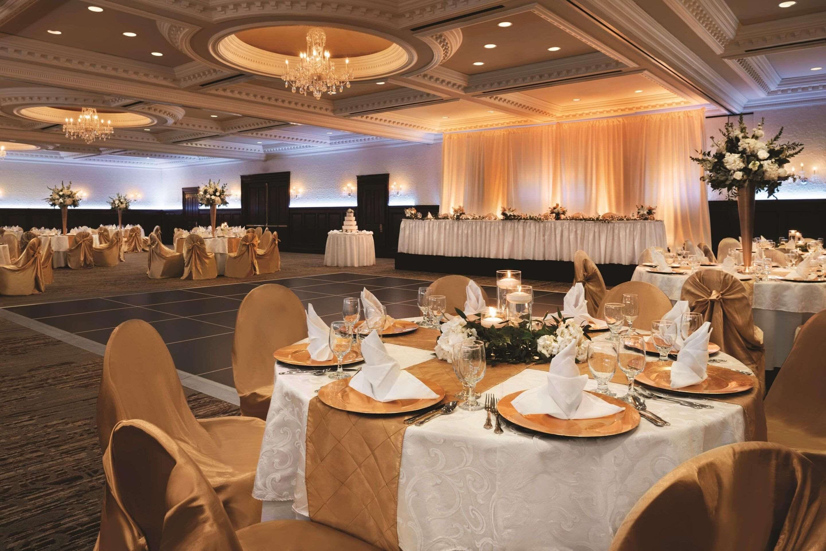 A banquet hall with round tables set for a formal event, featuring gold chair covers, white tablecloths, and elegant table settings.