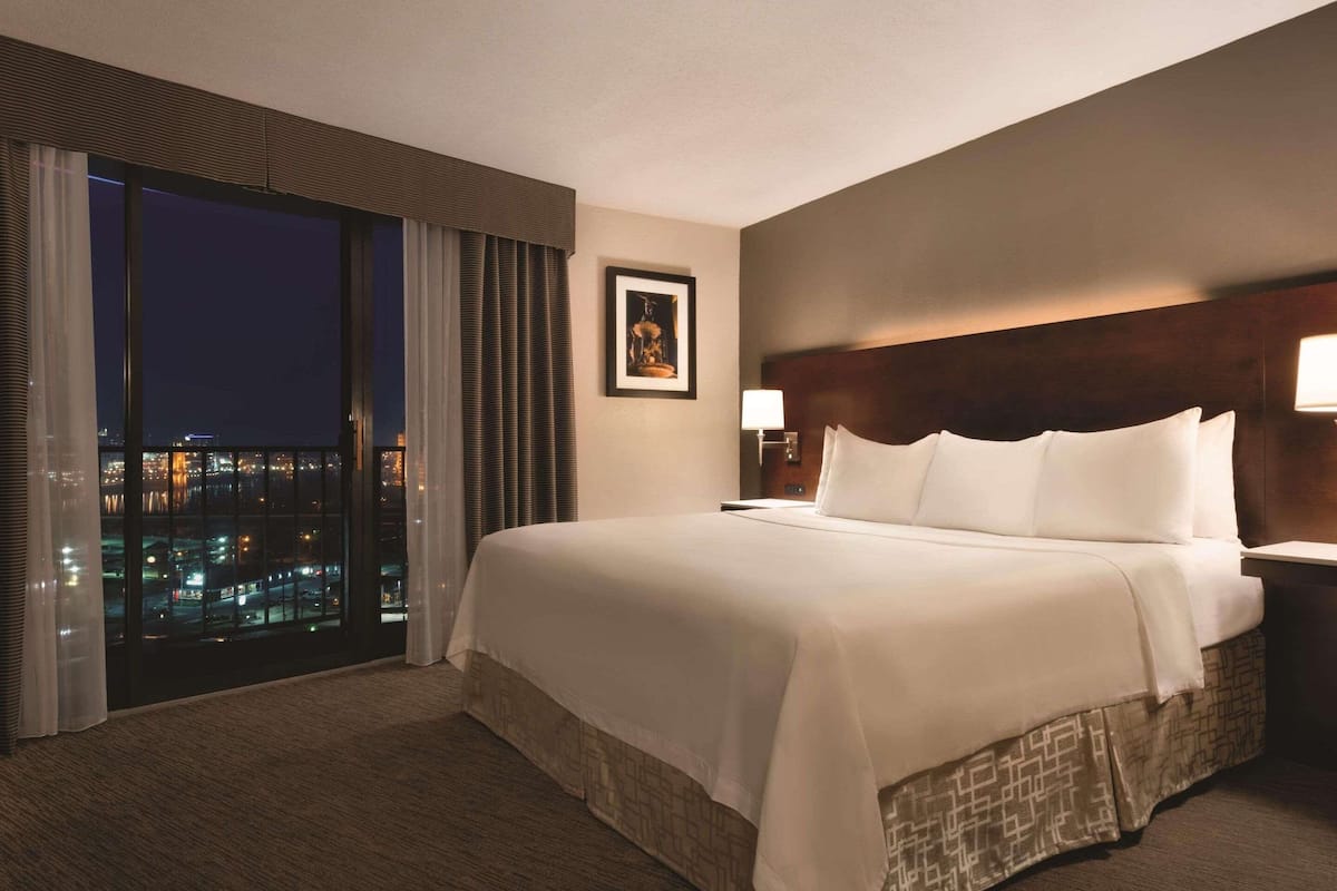 A hotel room with a large bed, a nightstand, a framed picture on the wall, and a view of the city at night through the window.