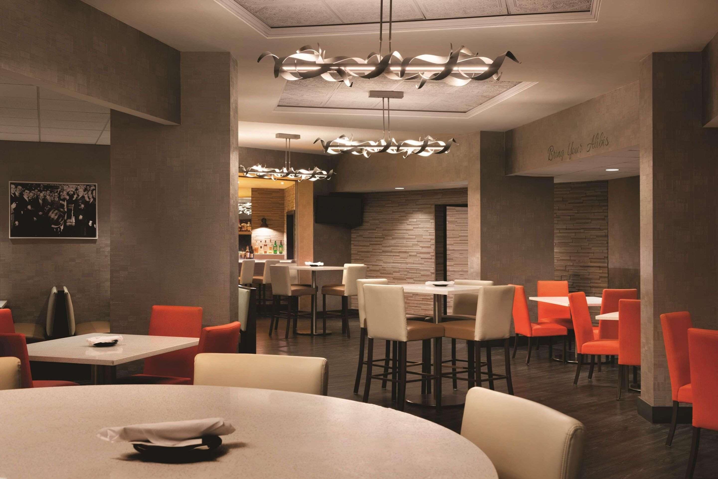 A restaurant with round tables and orange chairs, featuring a modern chandelier and a stone wall.