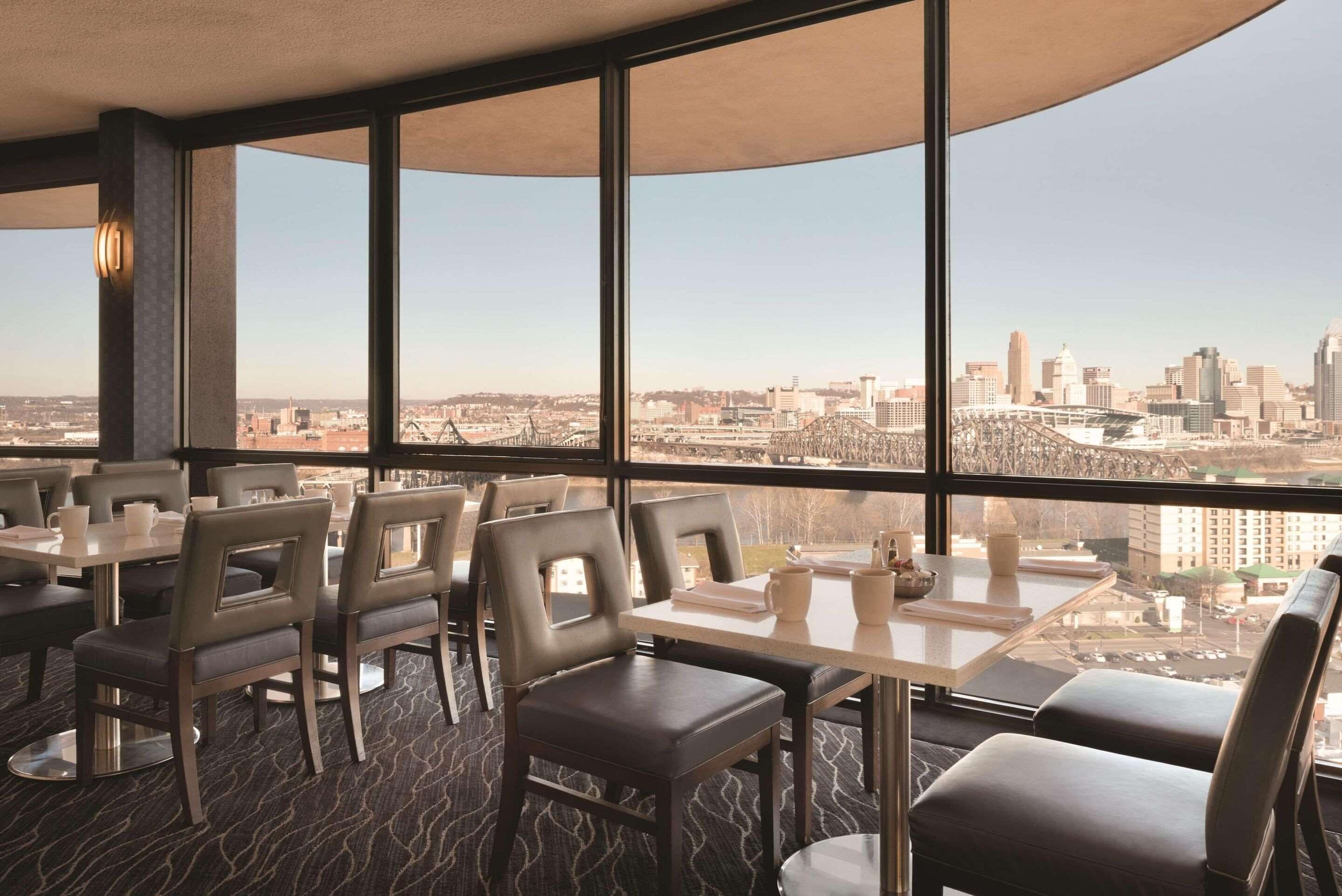 Un restaurante con amplios ventanales que ofrecen vistas a la ciudad. Las mesas están dispuestas con sillas, y el interior presenta un diseño moderno.