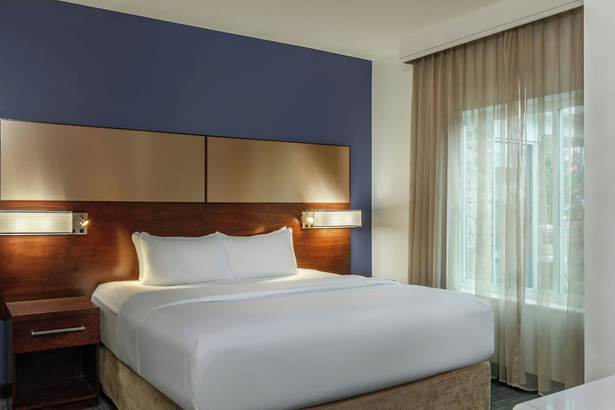 A hotel room with a large bed, a wooden headboard, bedside lamps, a nightstand, and a window with curtains.