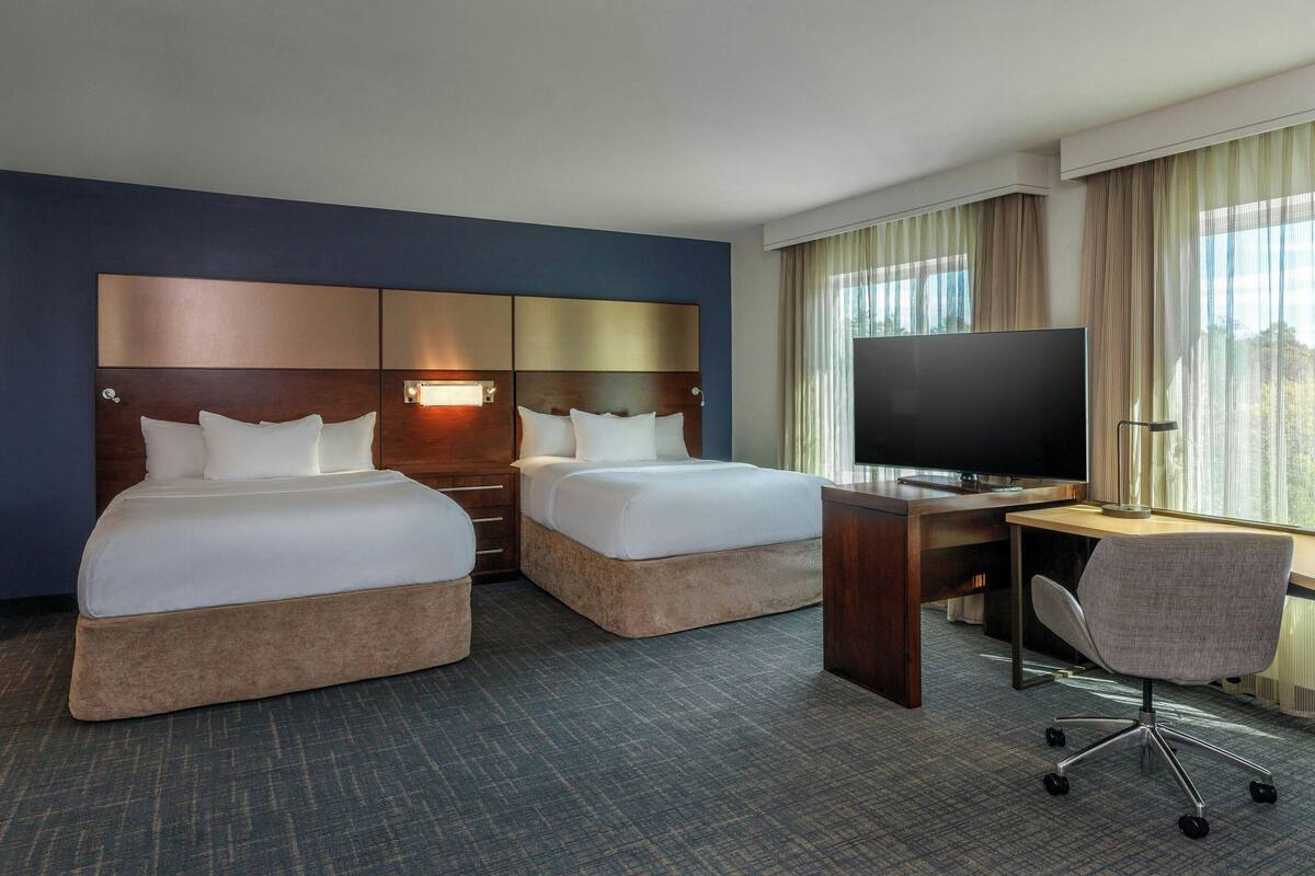 A hotel room with two beds, a desk, a chair, a TV, and large windows with curtains.