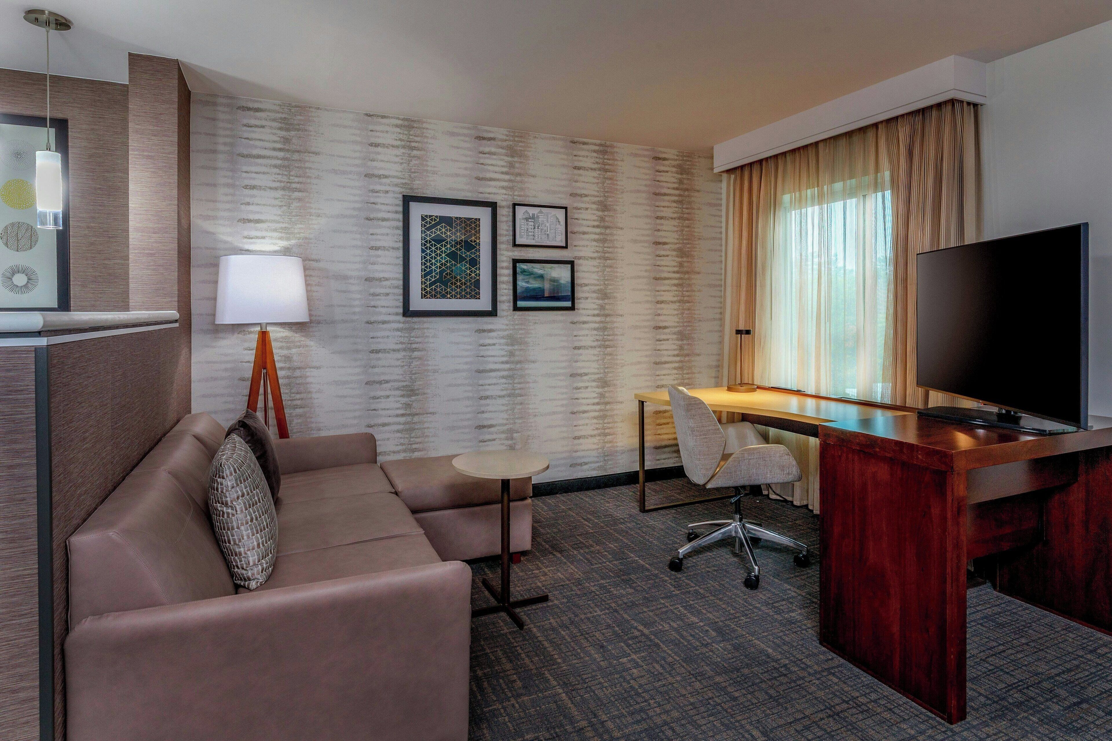 A hotel room with a sofa, a desk, a television, a lamp, and framed pictures on the wall.