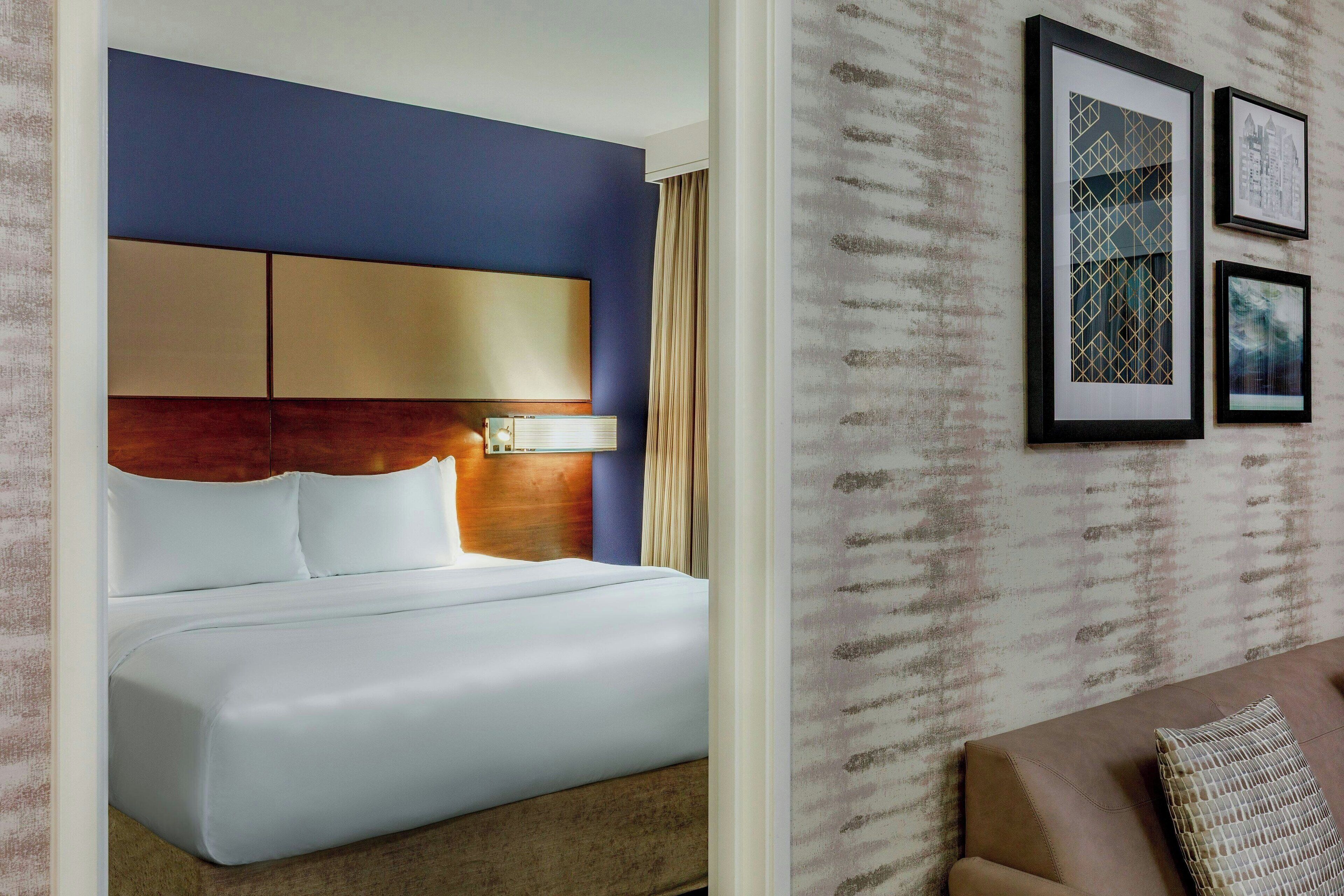 Ein Hotelzimmer mit einem großen Bett, einem Kopfteil mit einer Leuchte und einer Wand mit gerahmten Kunstwerken.