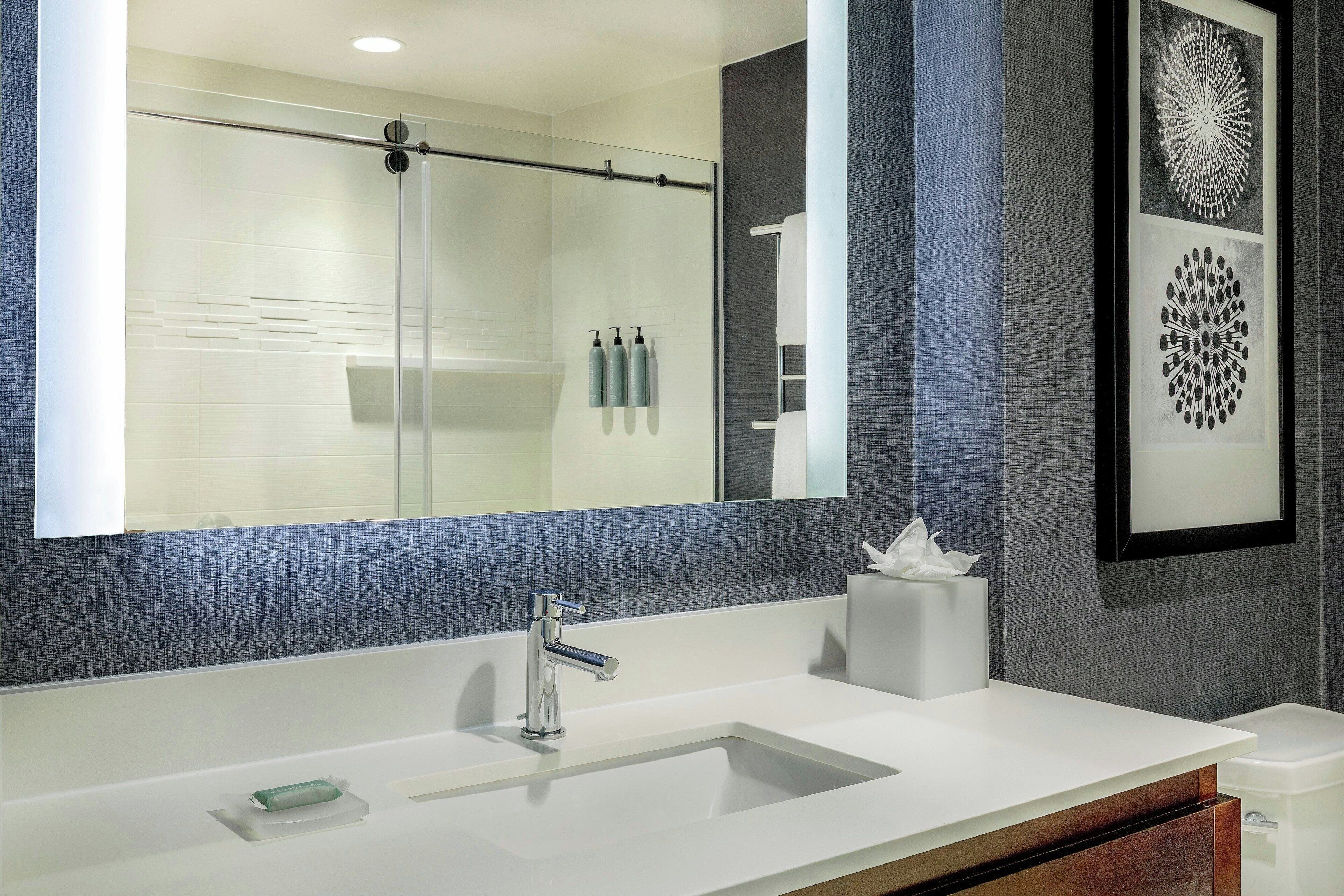 A modern bathroom with a large mirror, a glass shower enclosure, a white sink, and a towel dispenser.