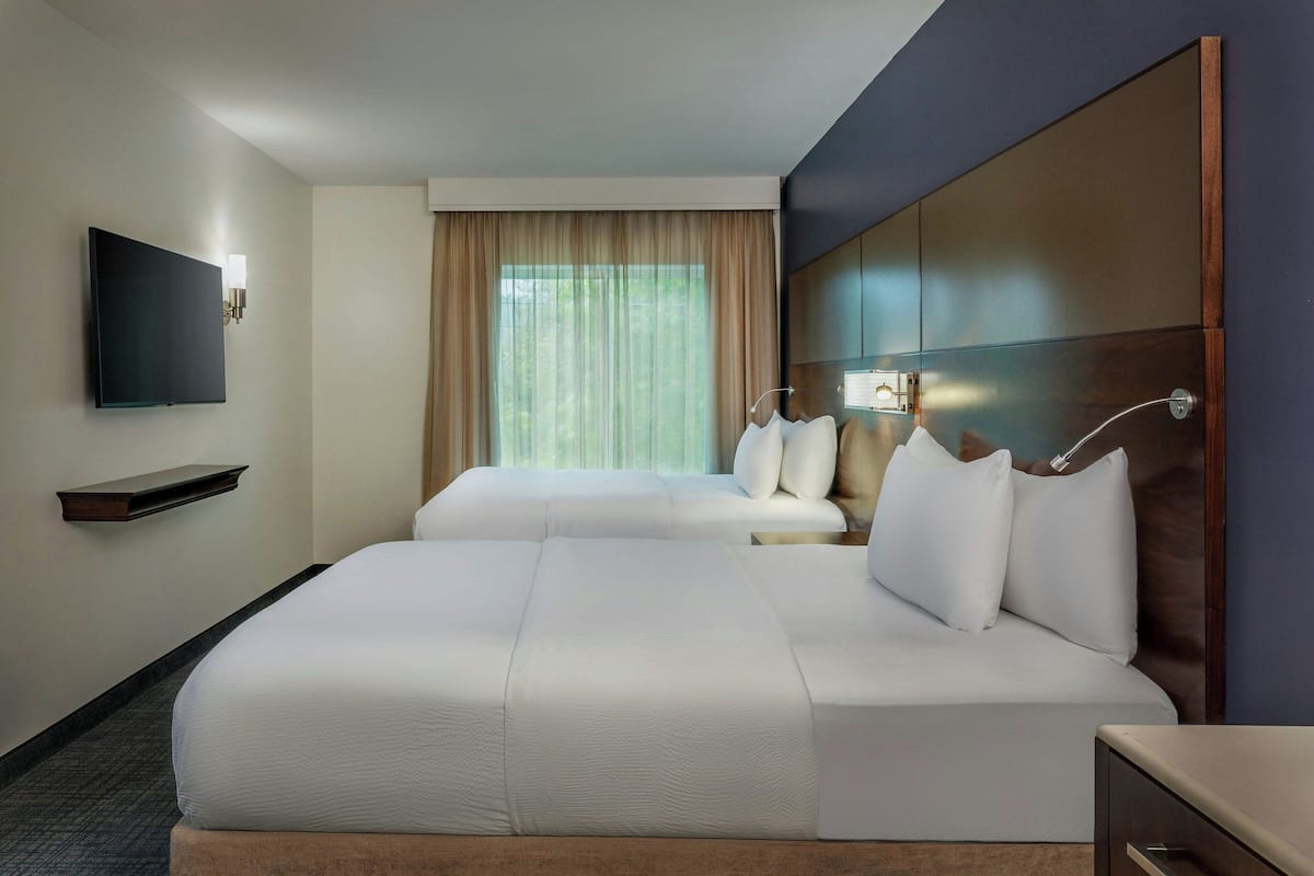 A hotel room with two beds, a flat-screen TV mounted on the wall, a bedside table with a lamp, and a window with curtains.