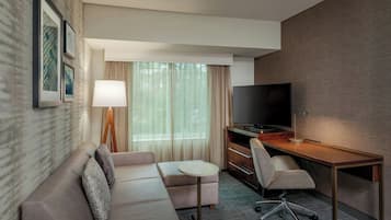 Una habitación de hotel moderna con un sofá, una pequeña mesa redonda, un escritorio con una silla, un televisor y una ventana con cortinas.