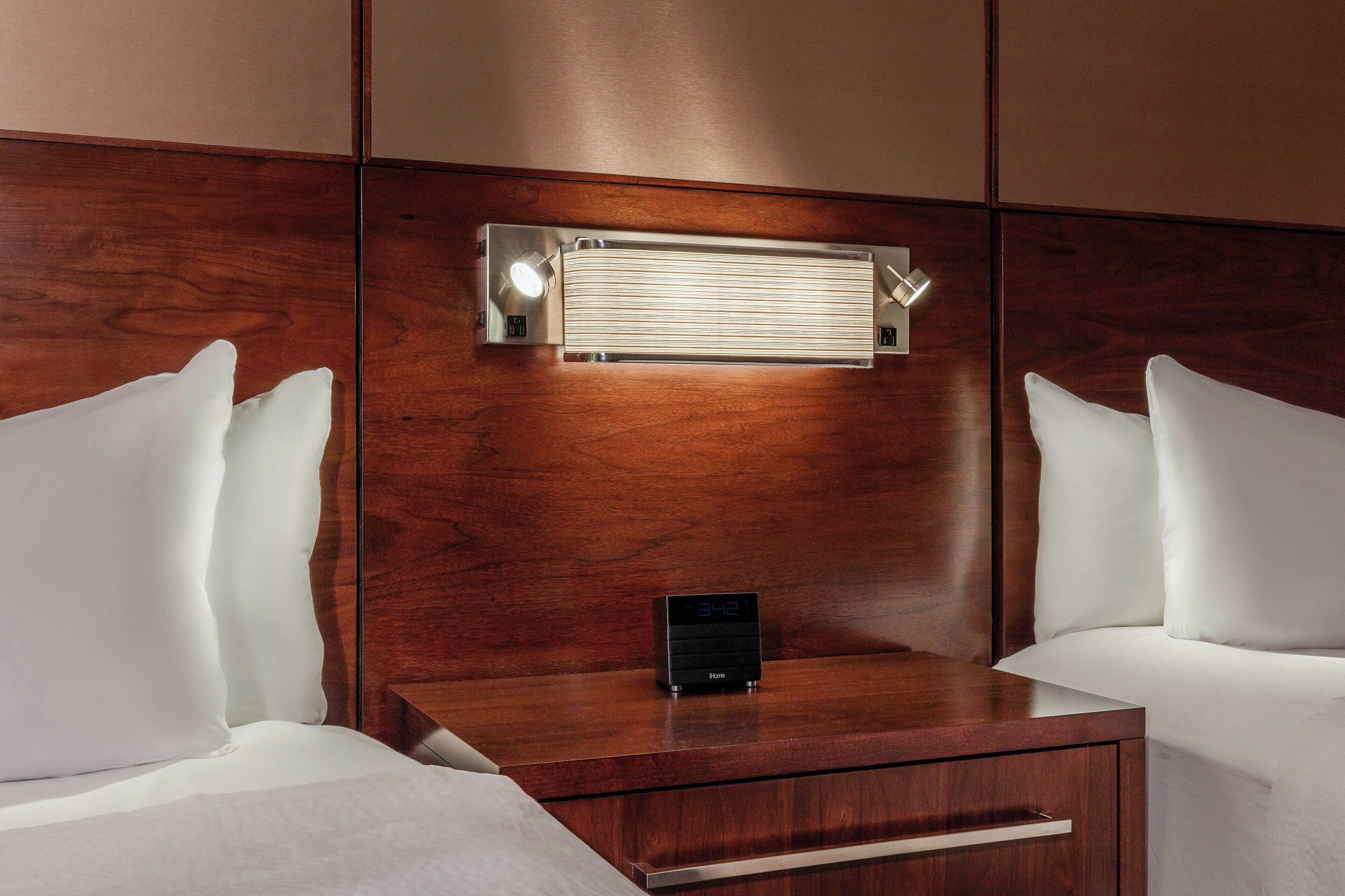A hotel room with a wooden headboard, a bedside table with a digital clock, and white bedding.