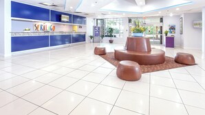 A reception area with a blue counter, a digital kiosk, and seating with a central planter. - Seralago Hotel and Suites Main Gate East (Kissimmee)